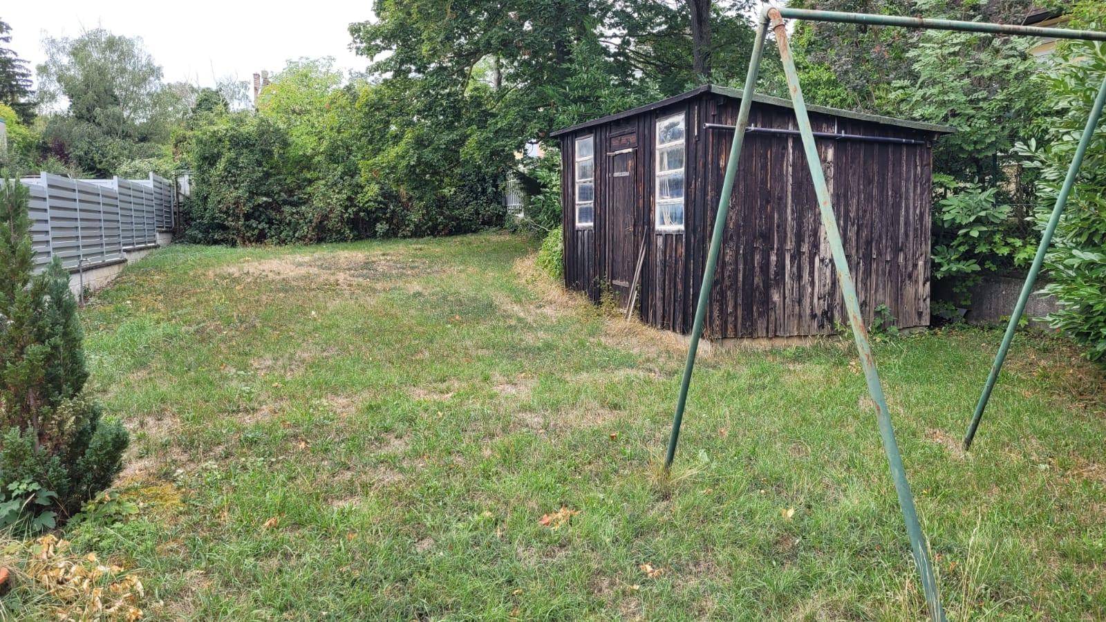 Rustikaler Holzschuppen in einem Gartenbereich mit Schaukelgestell und Metallzaun im Hintergrund.
