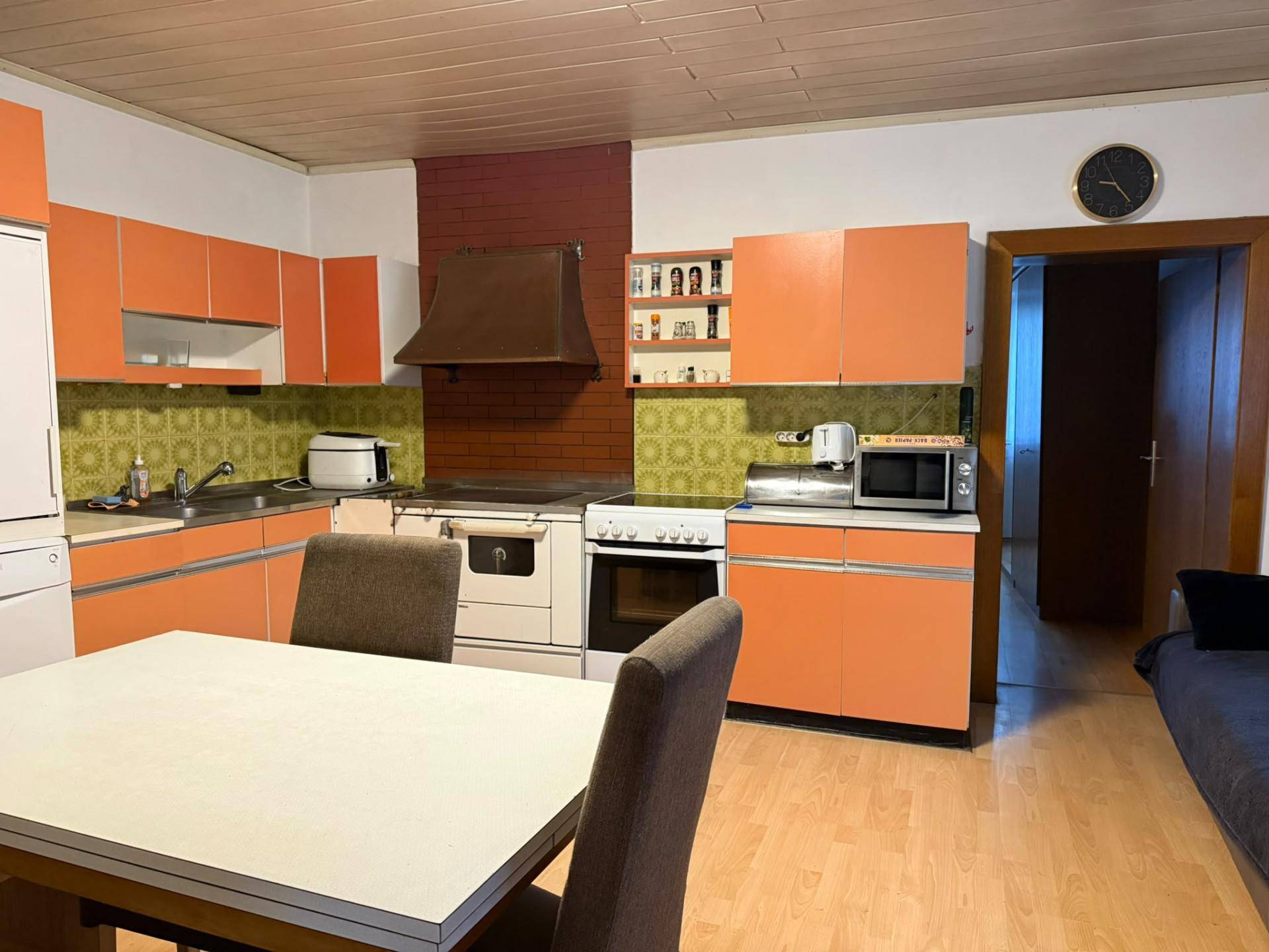 Küche im 80er-Jahre-Stil mit orangefarbenen Schränken, Esstisch und Holzfußboden, funktional eingerichtet.