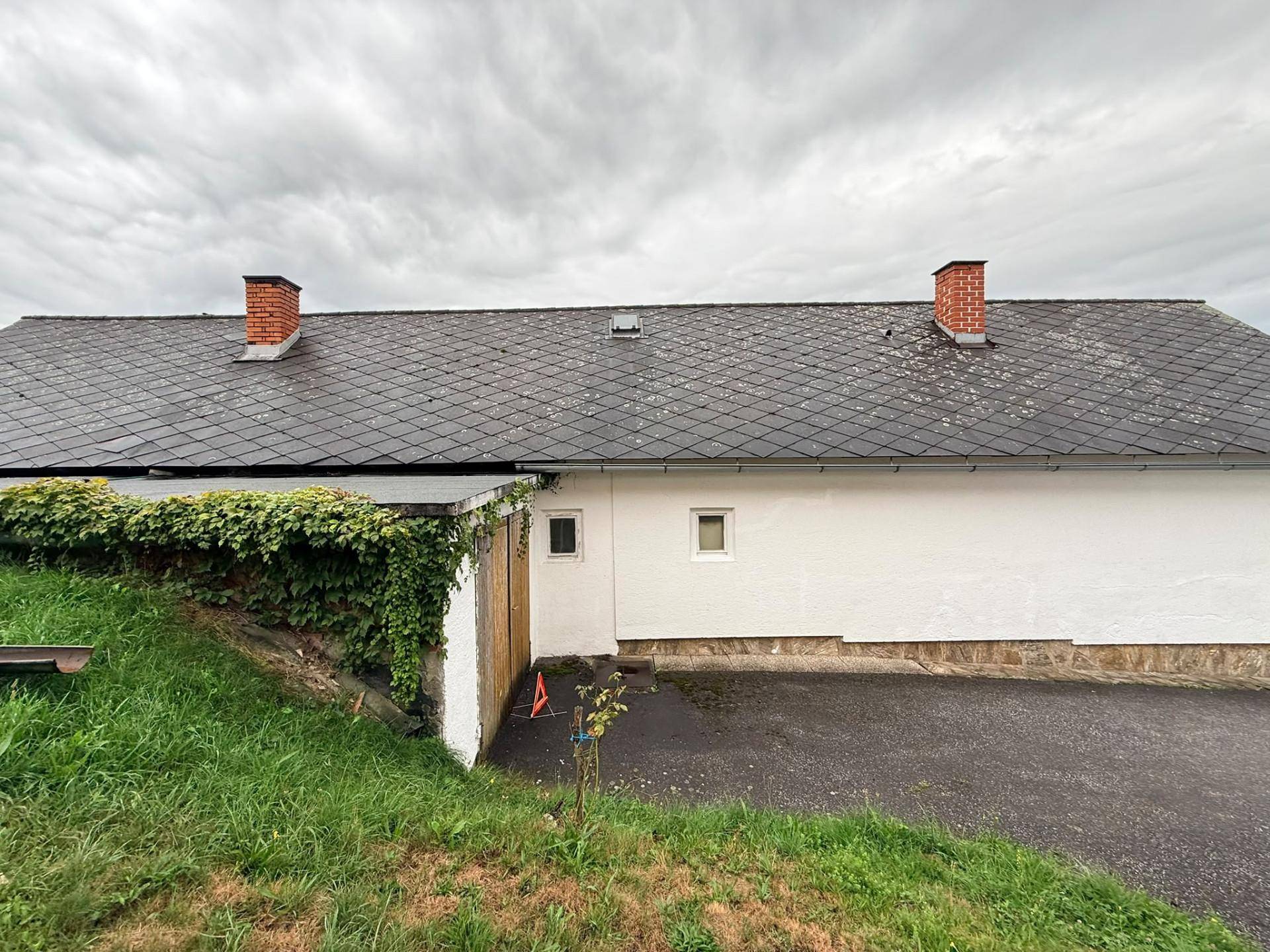 Seitenansicht des Gebäudes mit dunklem Dach, weißer Fassade und bewachsenem Anbau unter bewölktem Himmel.