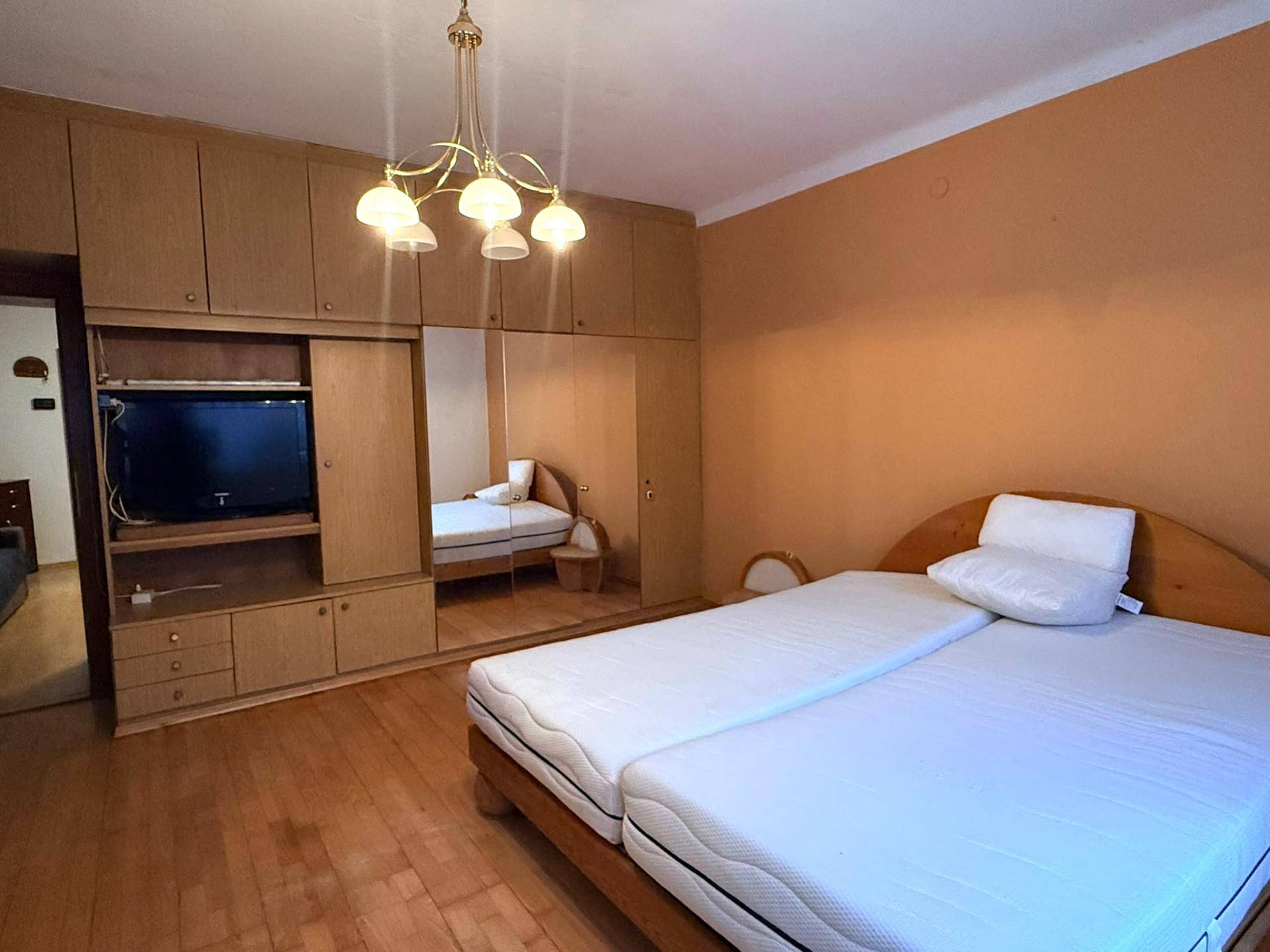 Schlafzimmer mit Doppelbett, großem Holzschrank und Spiegel, in warmen Farbtönen gehalten mit Parkettboden.