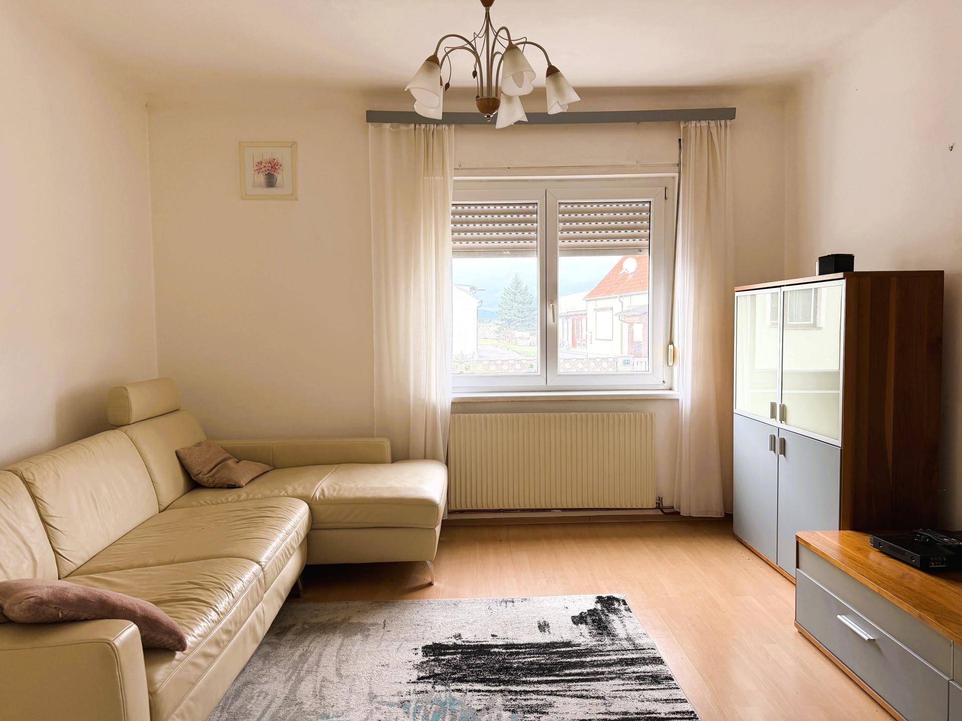 Gemütliches Wohnzimmer mit beigem Sofa, Holzboden und Blick ins Grüne durch das Fenster.