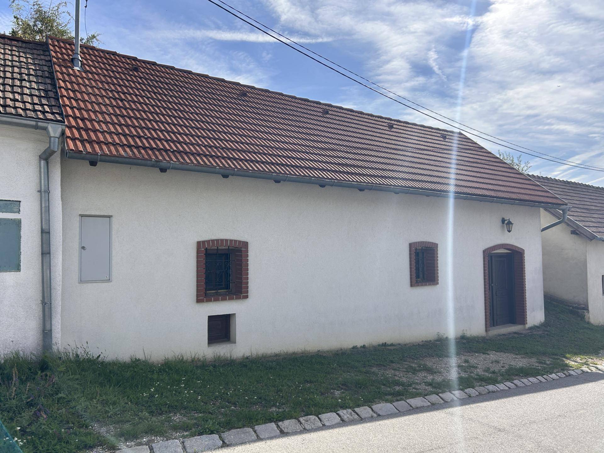 Längsansicht der weißen Fassade eines alten Gebäudes mit rotem Ziegeldach und kleinen, vergitterten Fenstern an einem sonnigen Tag.