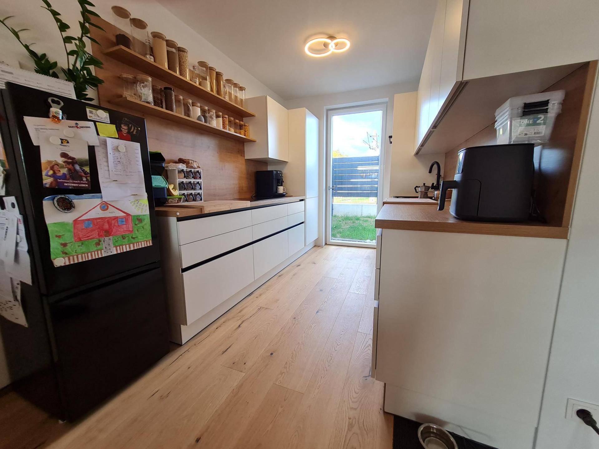 Helle Küche mit weißen Fronten, Holzregalen und Zugang zum Gartenbereich.