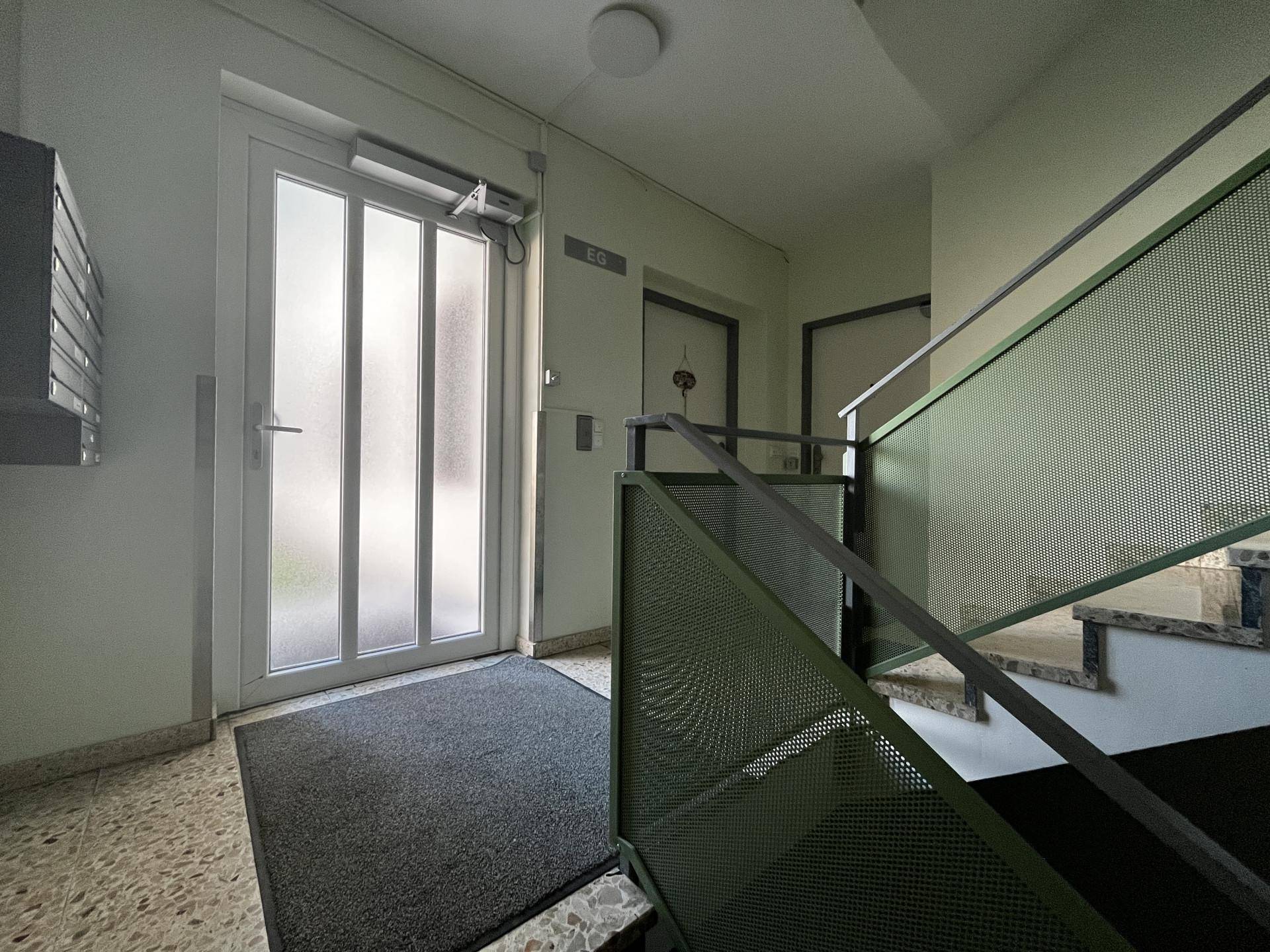 Eingangsbereich eines Mehrfamilienhauses mit Treppenhaus und Zugangstür zum Erdgeschoss.