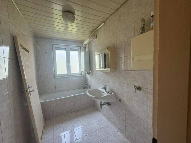 Einfaches Badezimmer mit Badewanne, Waschbecken und Fenster, gefliest in hellen Farbtönen.