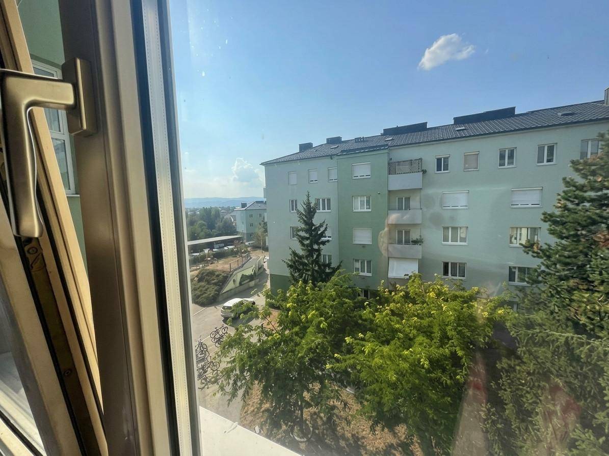 Blick aus dem Fenster auf ein gegenüberliegendes Wohngebäude und grüne Bäume.