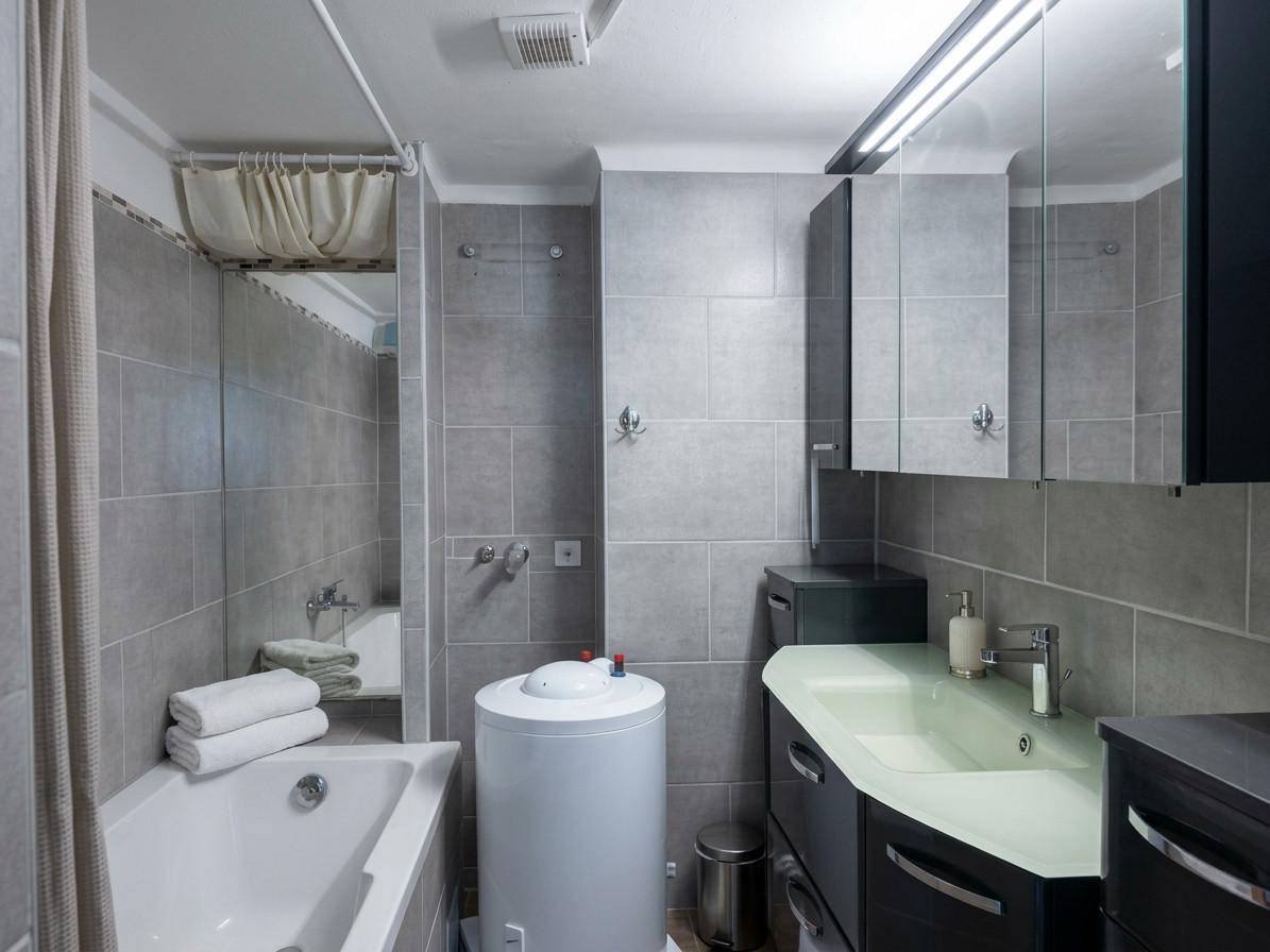 Badezimmer mit grauen Fliesen, Badewanne, Waschbecken und einem Warmwasserboiler.