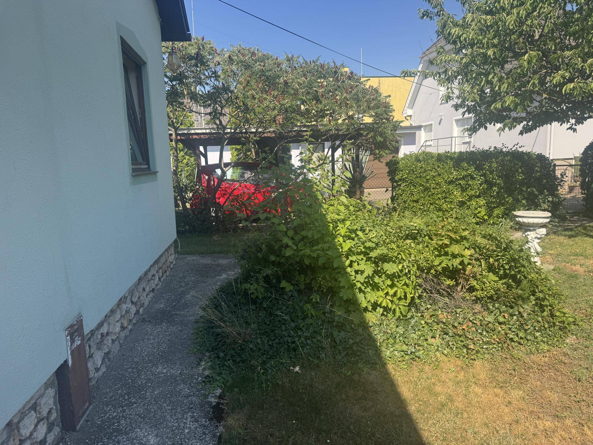 Seitenansicht des Hauses mit einem Weg, Büschen und einem roten Auto unter einem Carport.