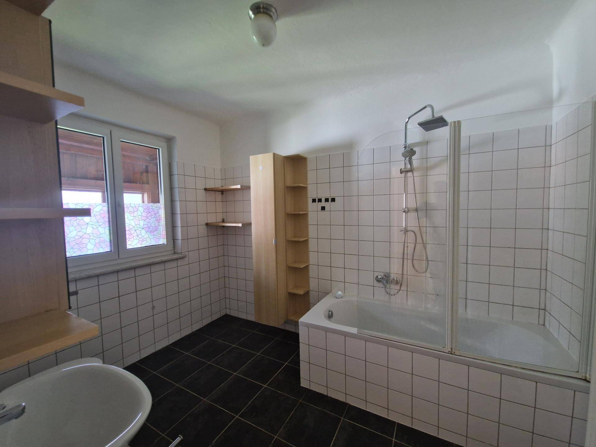 Gefliestes Badezimmer mit Badewanne, Dusche und Fenster, ausgestattet mit einfachen Regalen.