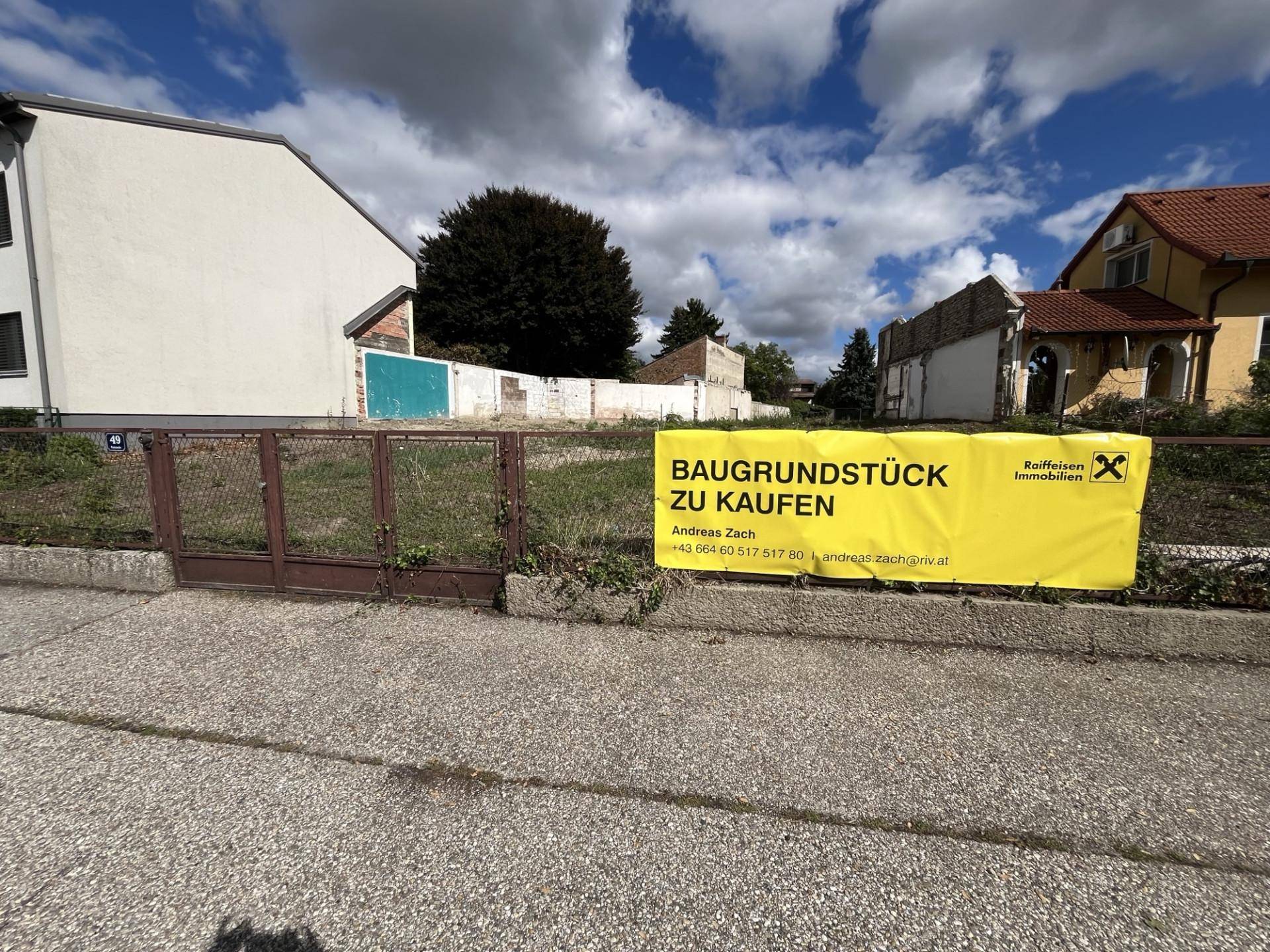 Baugrundstück mit Verkaufsbanner und umgebenden Gebäuden unter bewölktem Himmel.