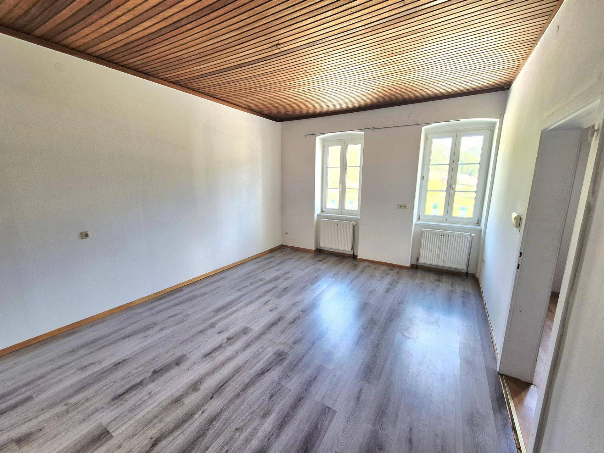 Helles Wohnzimmer mit Holzbalkendecke, dunklem Laminatboden und zwei Fenstern.