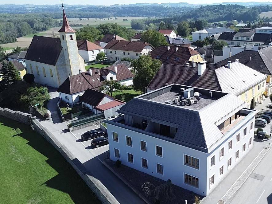 Luftaufnahme eines Dorfes mit Kirche, Wohngebäuden und weitem Blick über die grüne Landschaft.