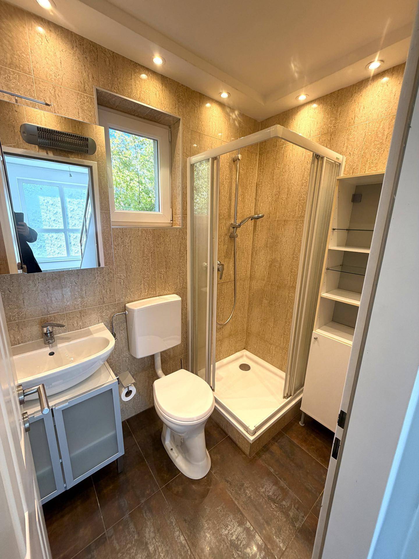 Modernes Badezimmer mit beigen Fliesen, Duschkabine, WC, Waschbecken und einem Fenster für Tageslicht.