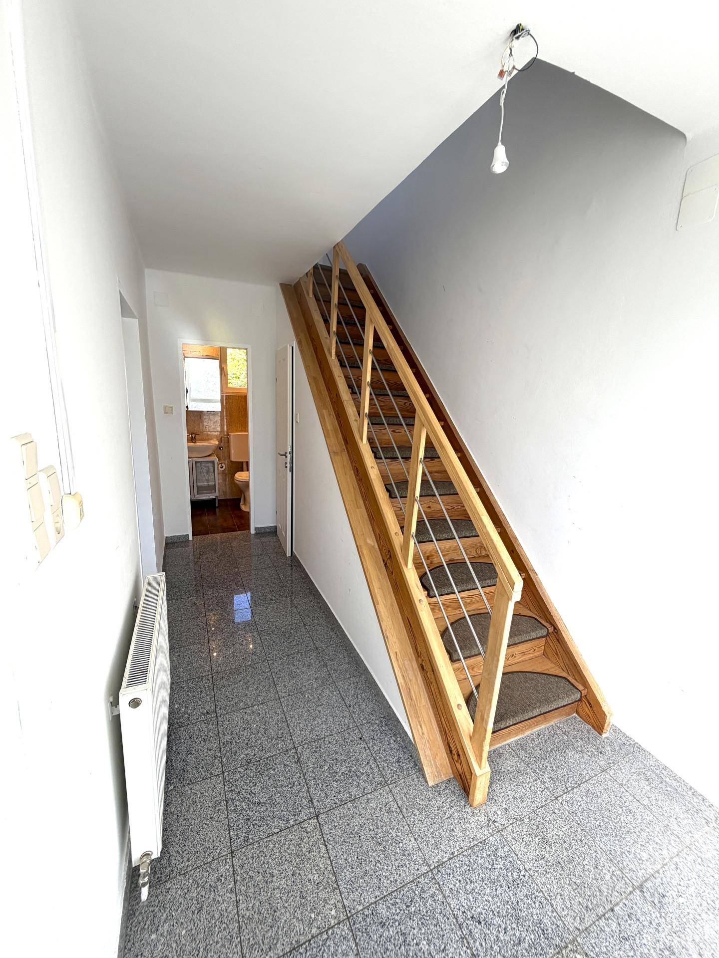 Heller Flur mit grauen Fliesen und einer Holztreppe, die in das obere Stockwerk führt.