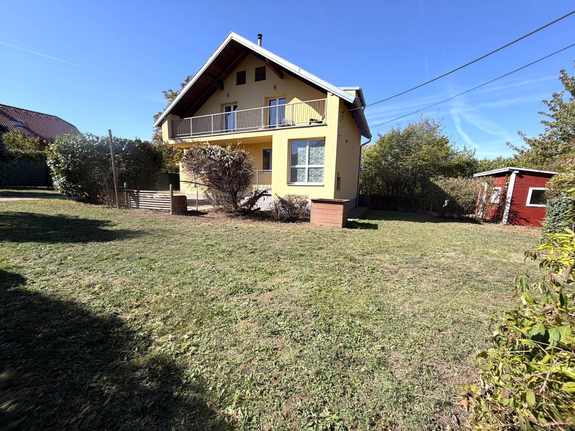 Ansicht des gelben Einfamilienhauses von der Gartenseite mit großem Rasen und einem kleinen Geräteschuppen.