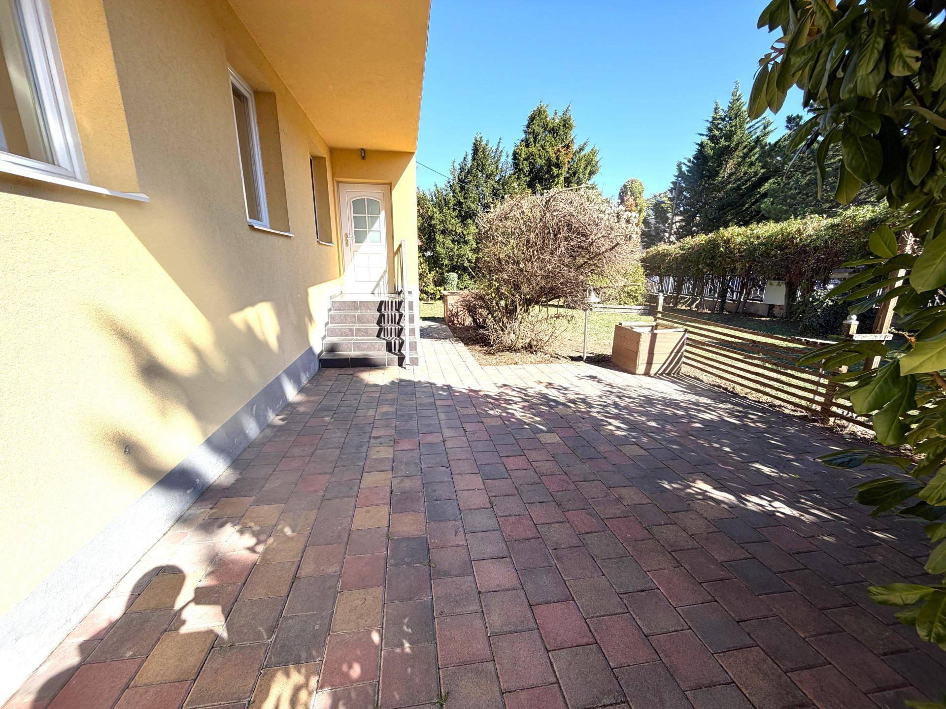Gepflasterte Terrasse neben dem Haus mit Zugang zur Eingangstür, umgeben von Grünflächen.