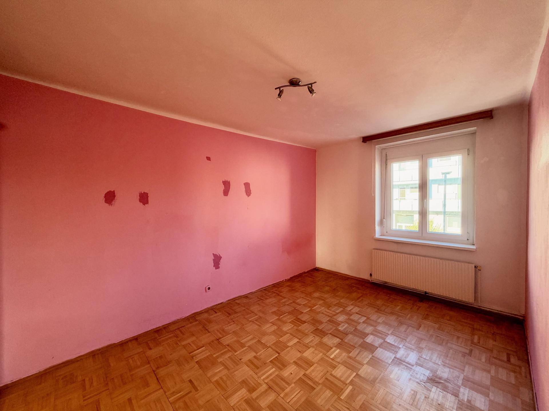 Schlafzimmer mit rosa Wänden, Parkettboden und einem Fenster, das viel Tageslicht hereinlässt.