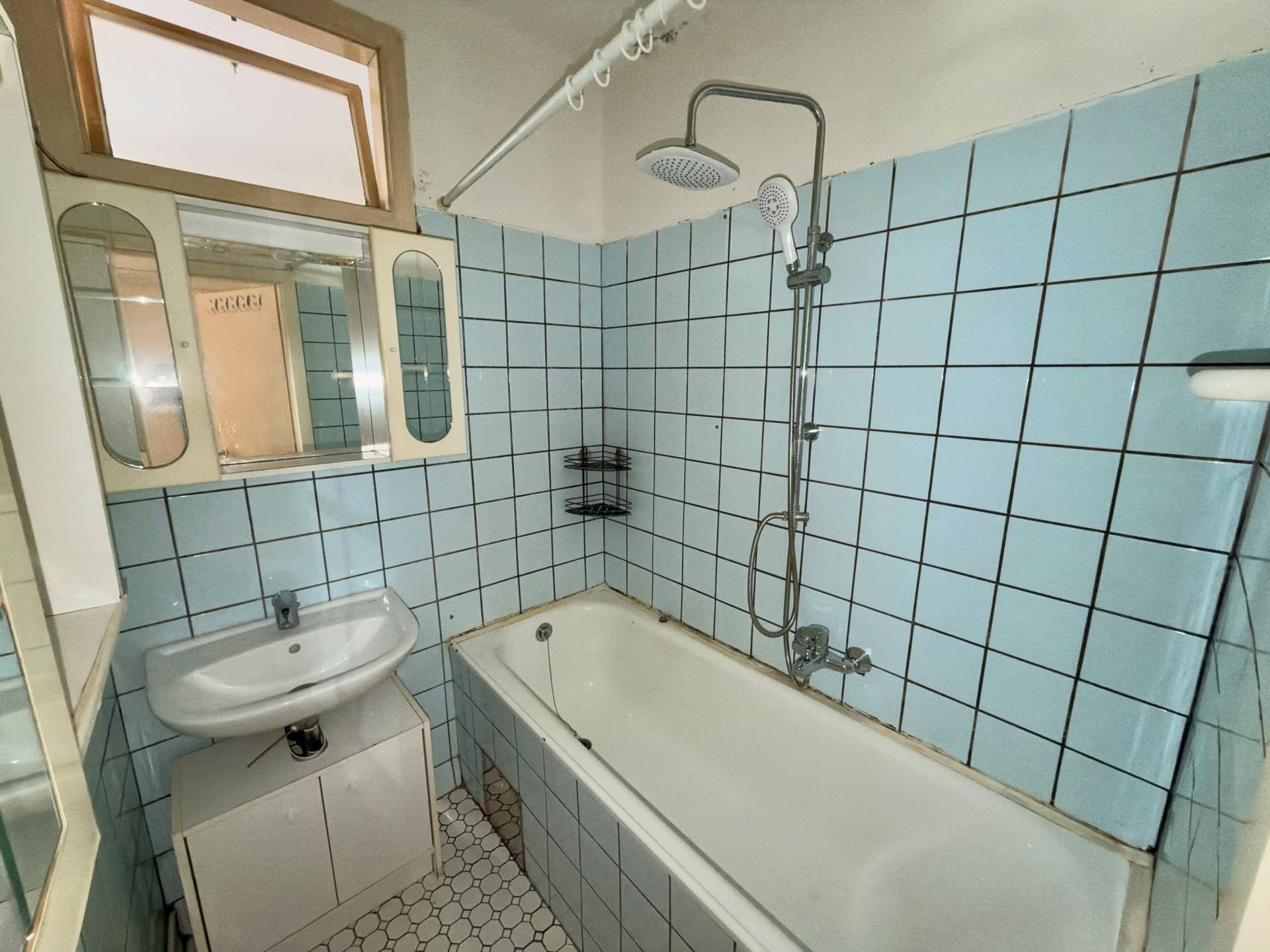 Badezimmer mit hellblauen Fliesen, Badewanne, Dusche und einem kleinen Fenster.