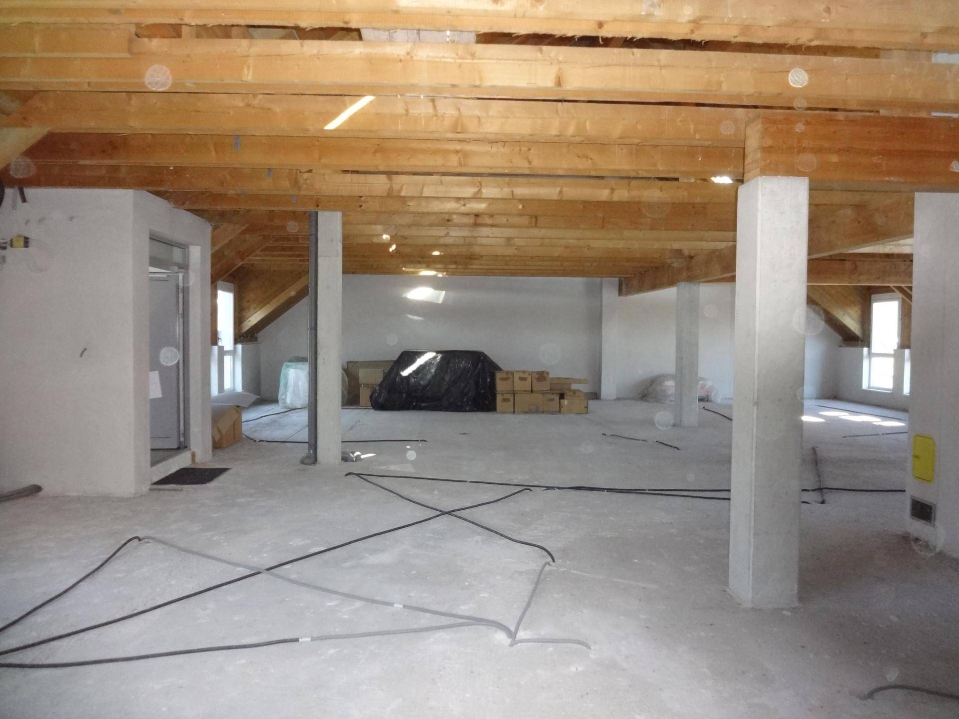 Unfertiger Dachbodenbereich mit freiliegenden Holzbalken, Betonboden und Säulen, der auf den Ausbau wartet.