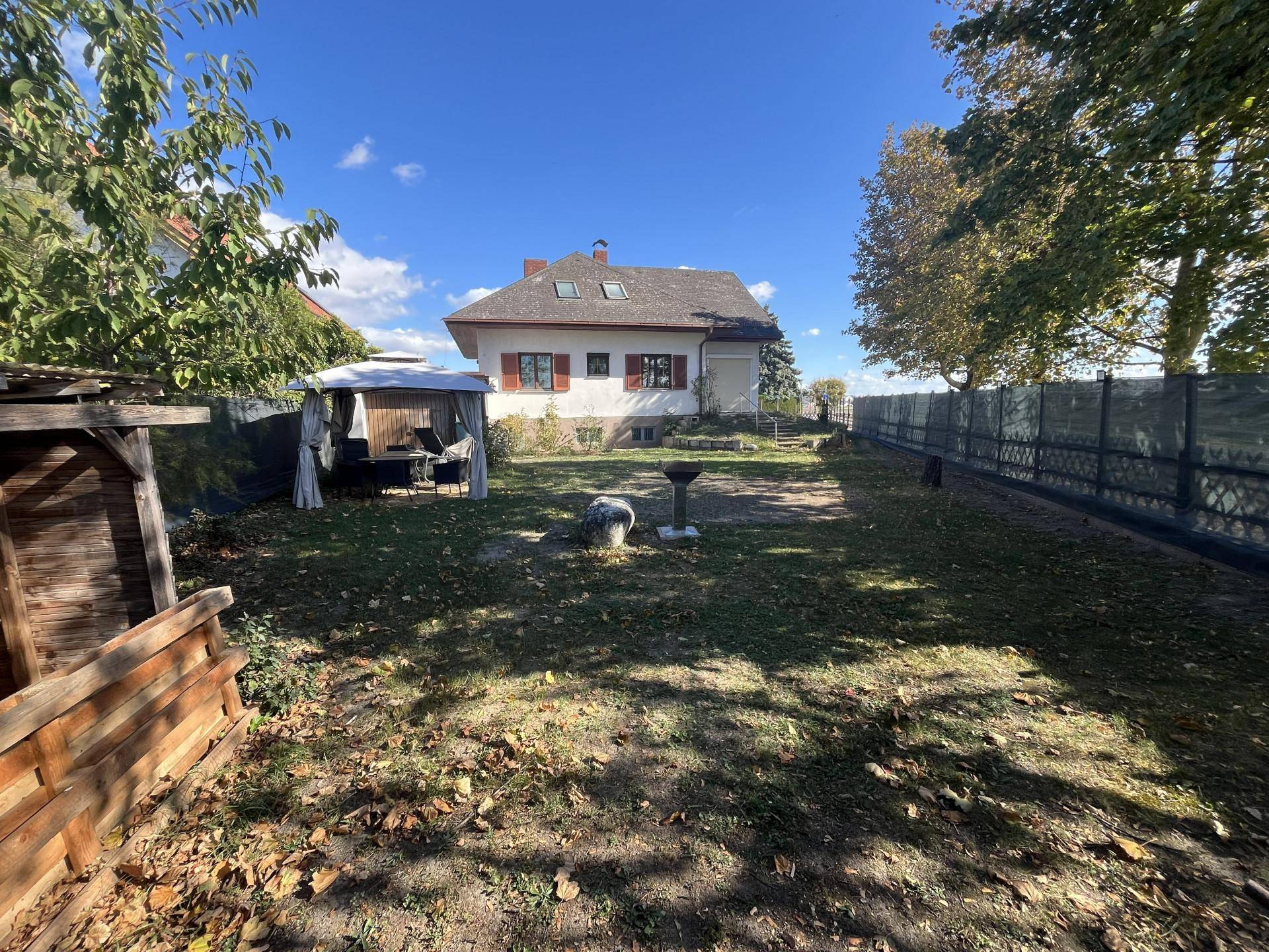 Weitläufiger Gartenbereich mit dem Haus im Hintergrund, Grillplatz und schattigem Sitzbereich.