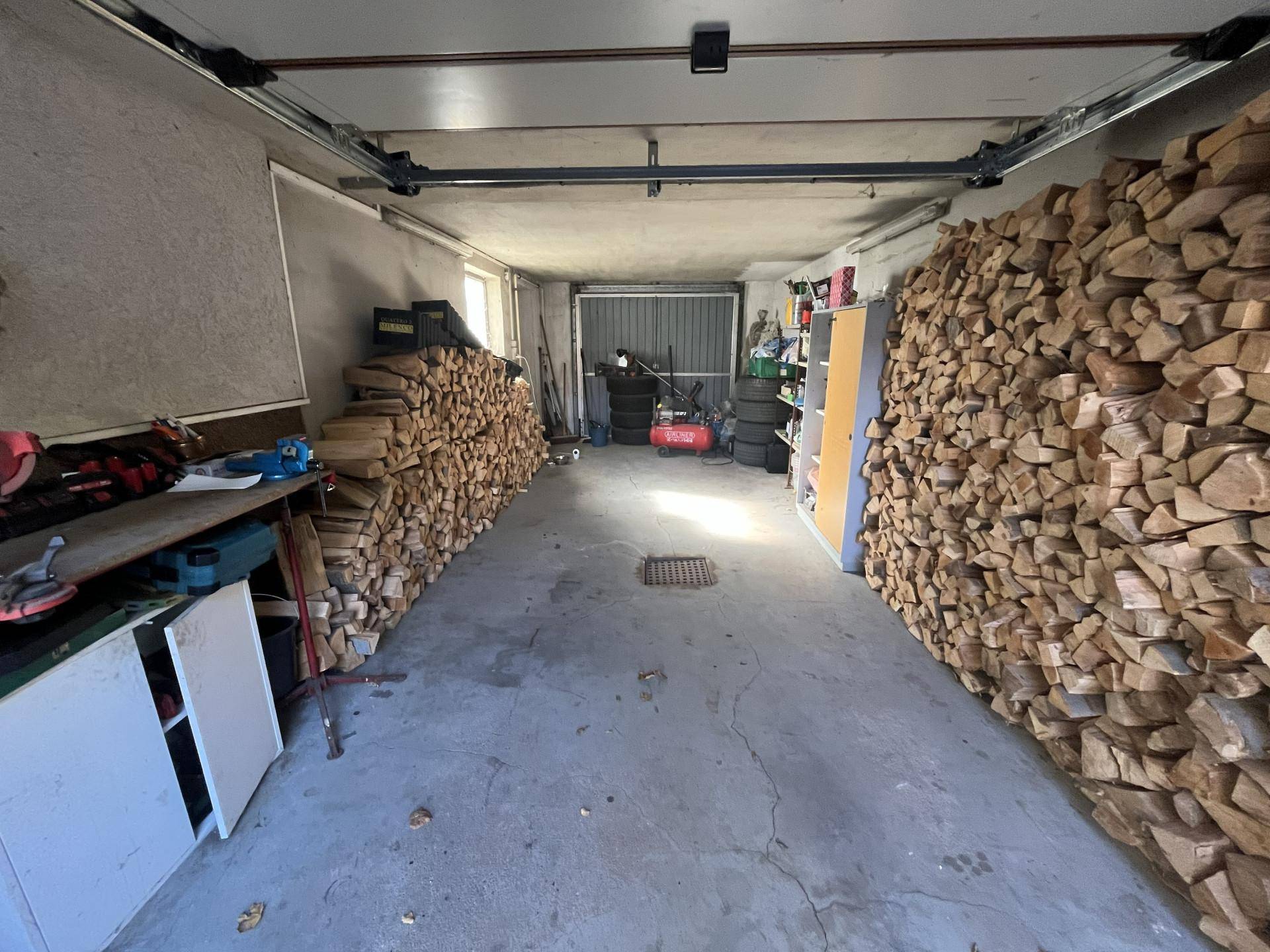 Geräumige Garage mit Betonboden, viel Stauraum und großen Stapeln Brennholz an den Wänden.