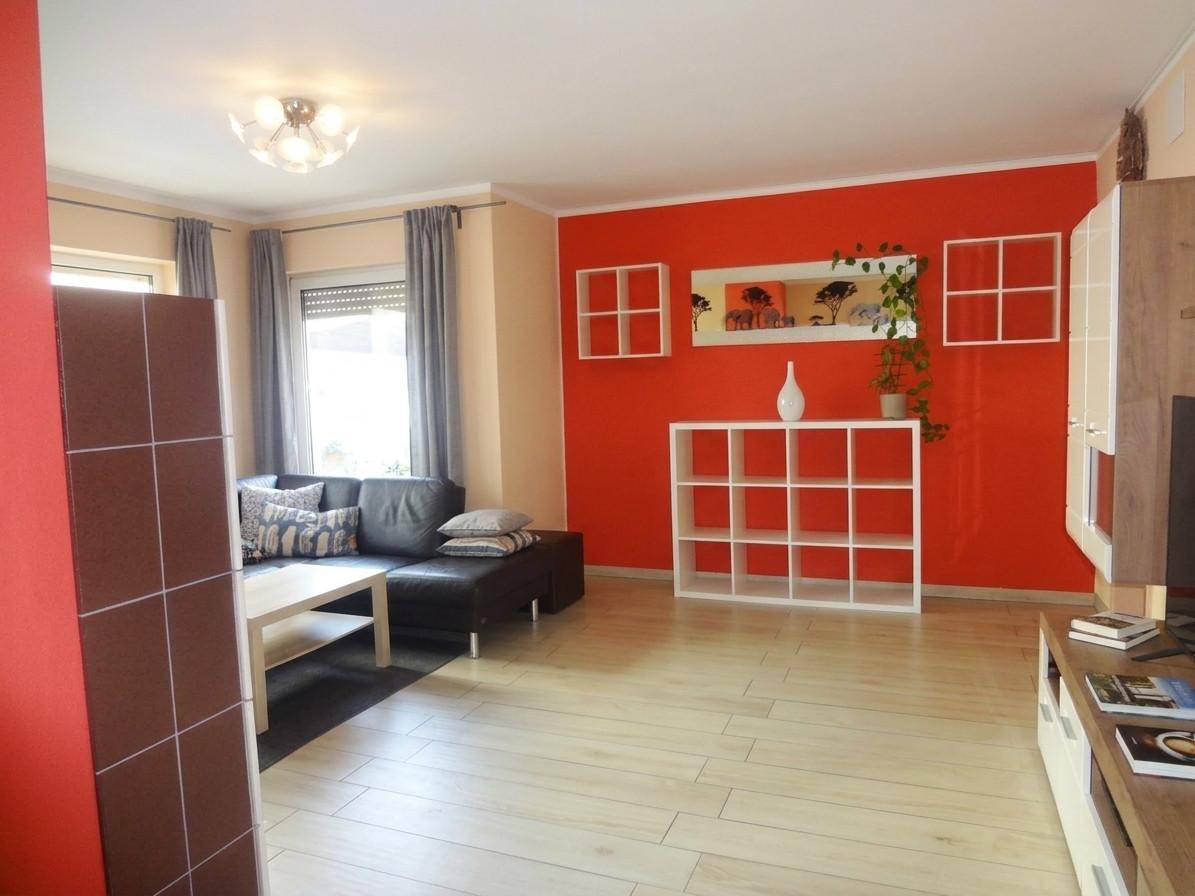 Helles Wohnzimmer mit roter Akzentwand, hellem Boden und moderner Einrichtung.