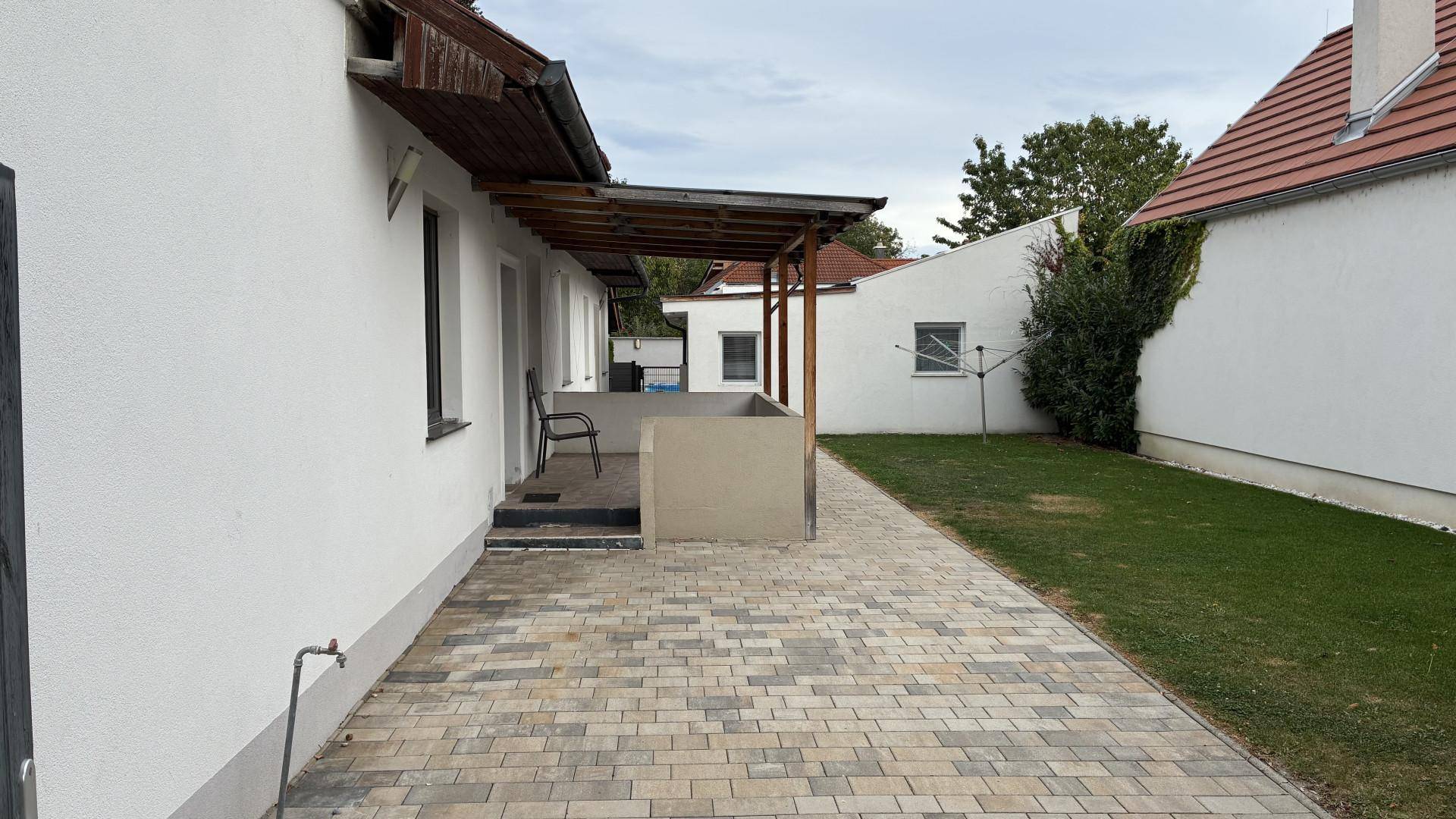 Langer gepflasterter Terrassenweg entlang des Hauses, der zu einem Rasenbereich führt.