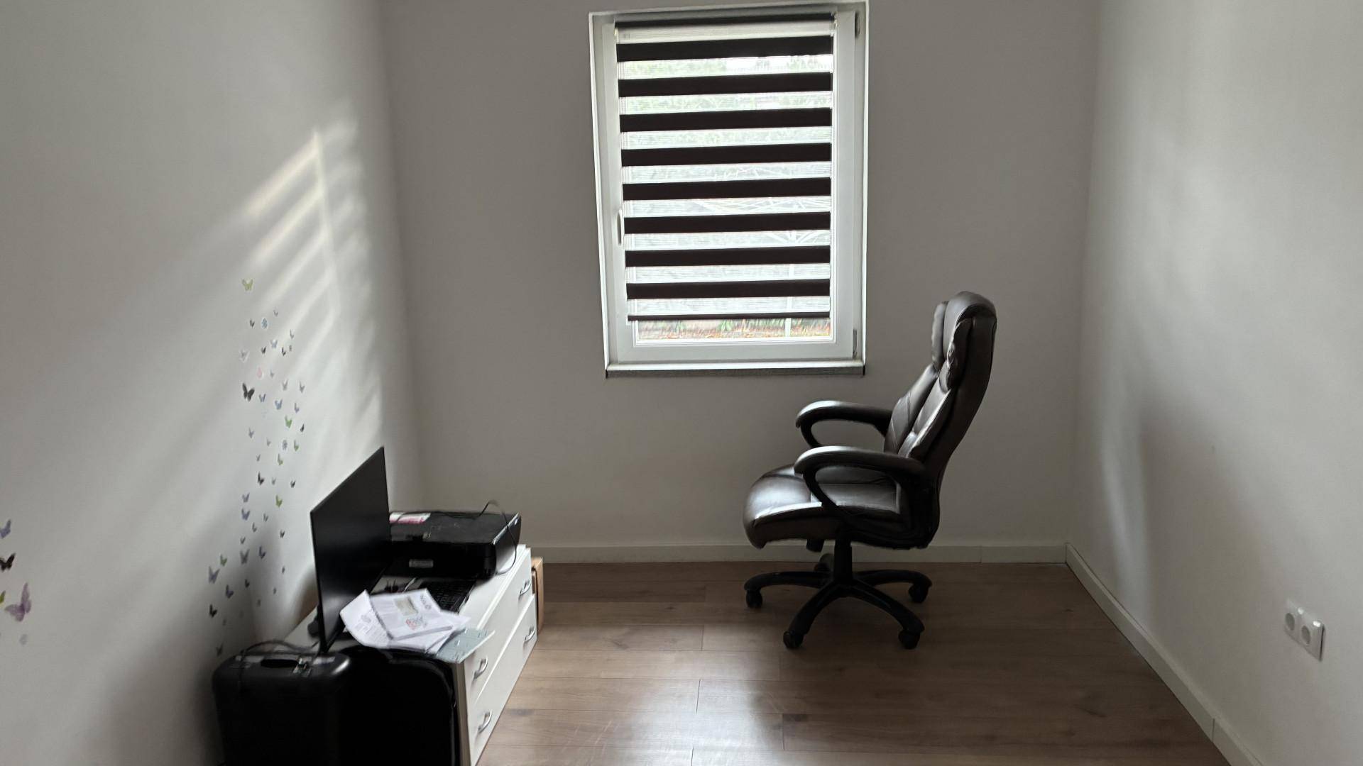 Kleines Arbeitszimmer mit Holzboden, einem Bürostuhl und einem Fenster mit Jalousien.