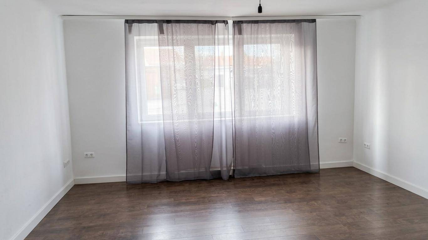 Geräumiges, leeres Zimmer mit dunklem Holzboden und großen Fenstern mit Vorhängen.