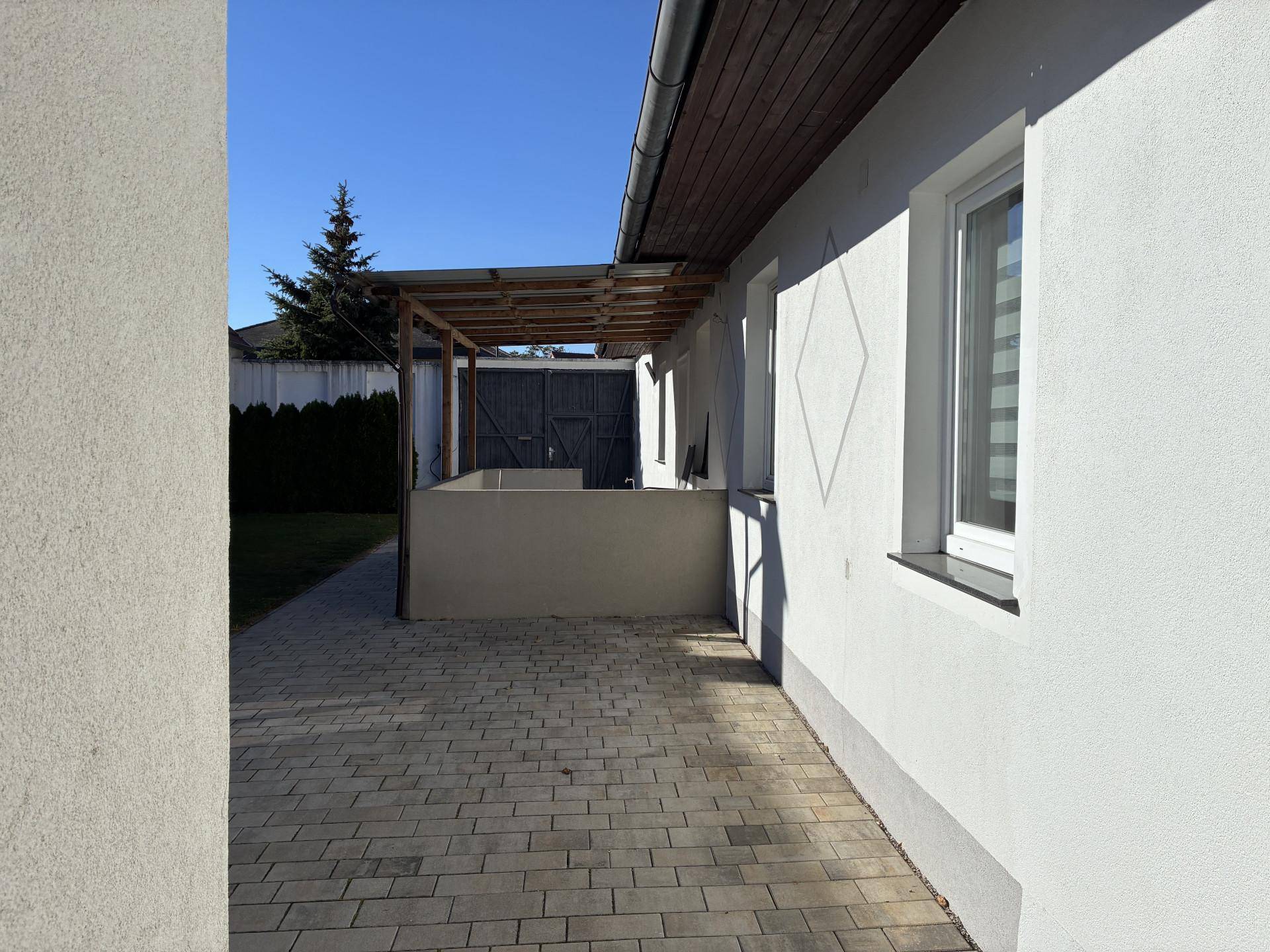 Sonnige gepflasterte Fläche neben dem Haus mit einer einfachen Holzüberdachung.