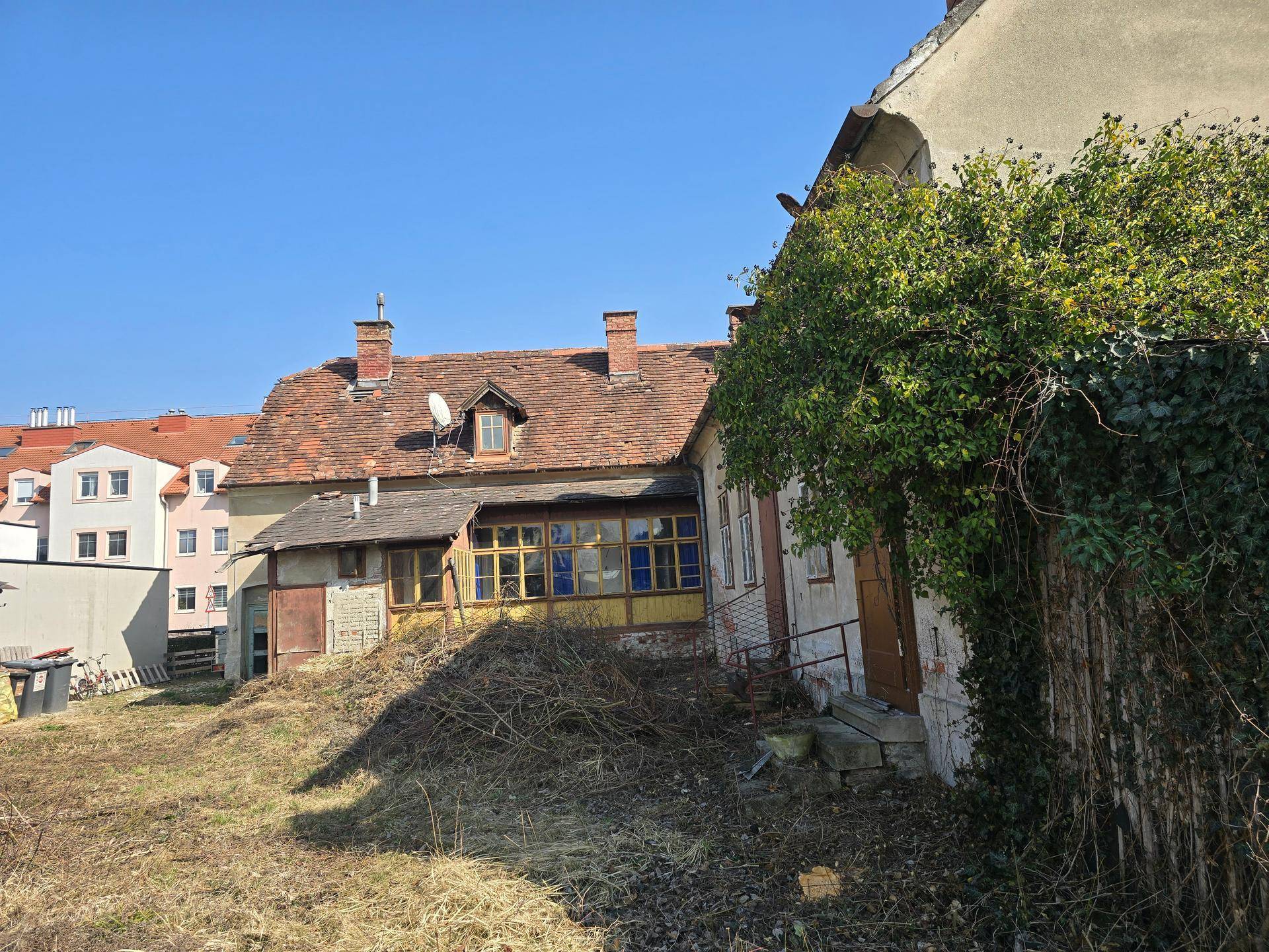 Rückansicht eines alten Wohnhauses mit Anbau, umgeben von einem verwilderten Gartenbereich.