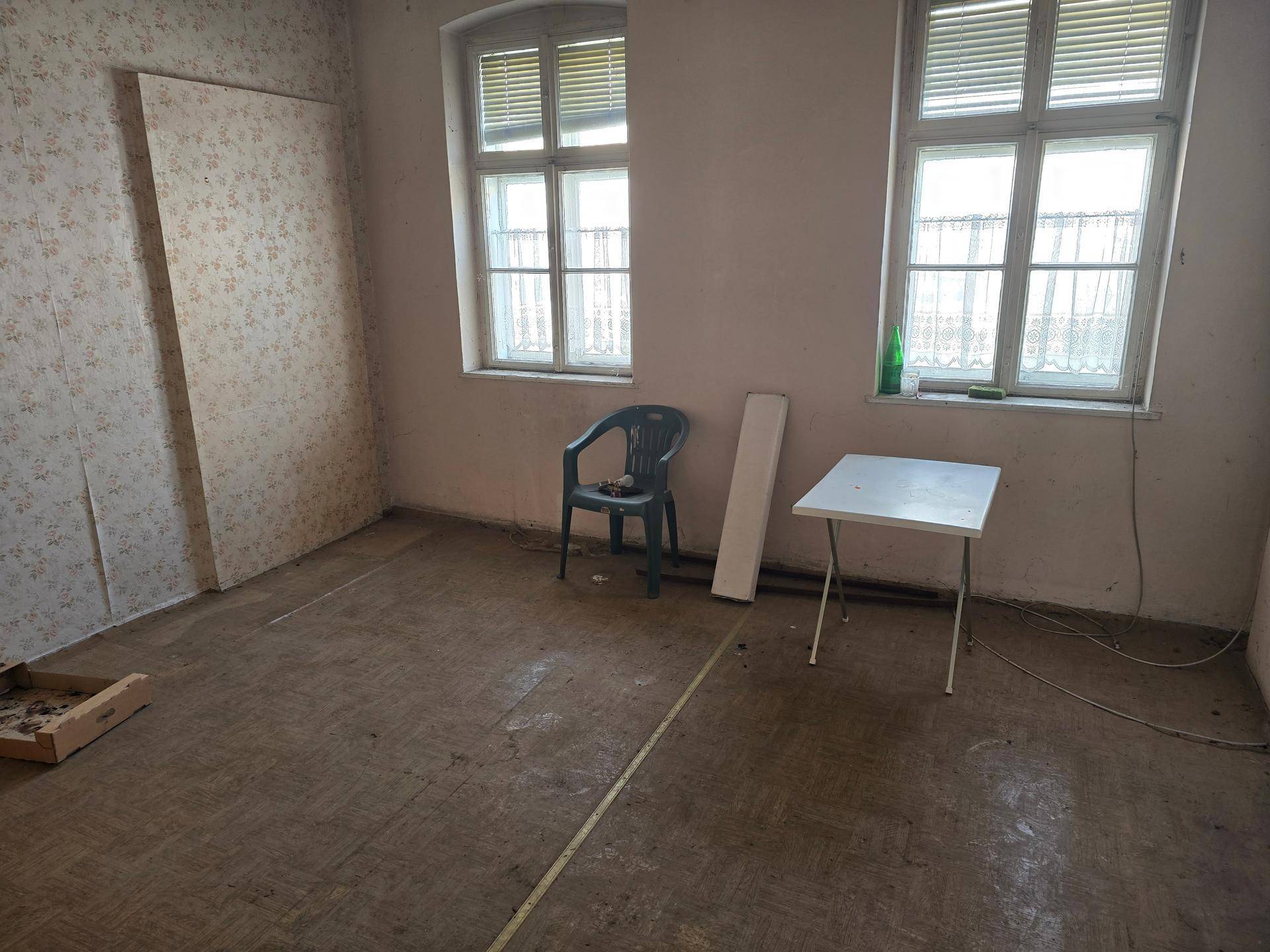 Leerer Raum mit zwei Fenstern, abgenutztem Boden und Tapetenresten, bereit zur Neugestaltung.
