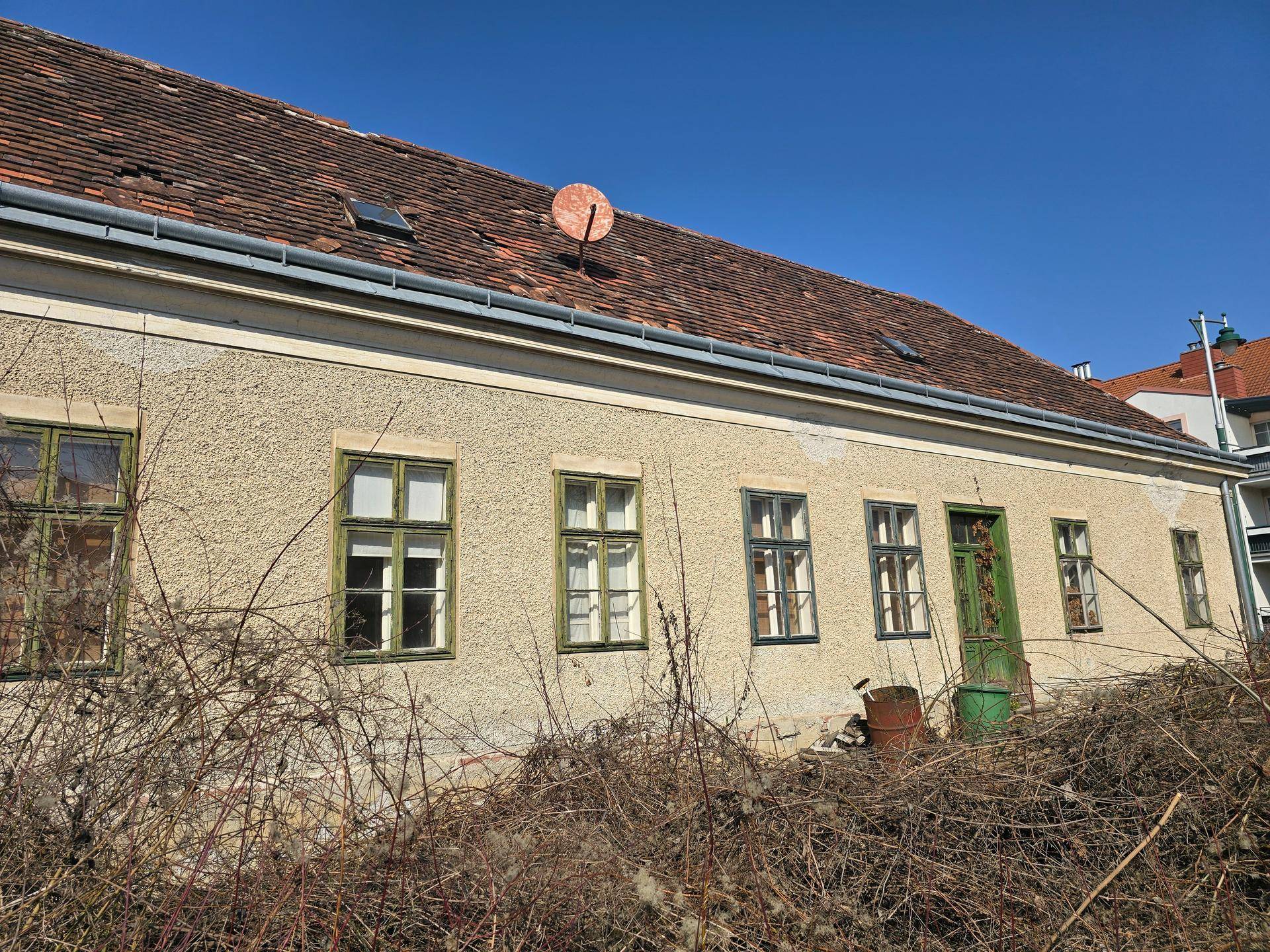 Seitenansicht des Wohnhauses mit bröckelndem Putz, alten Fenstern und einem verwilderten Grundstück.