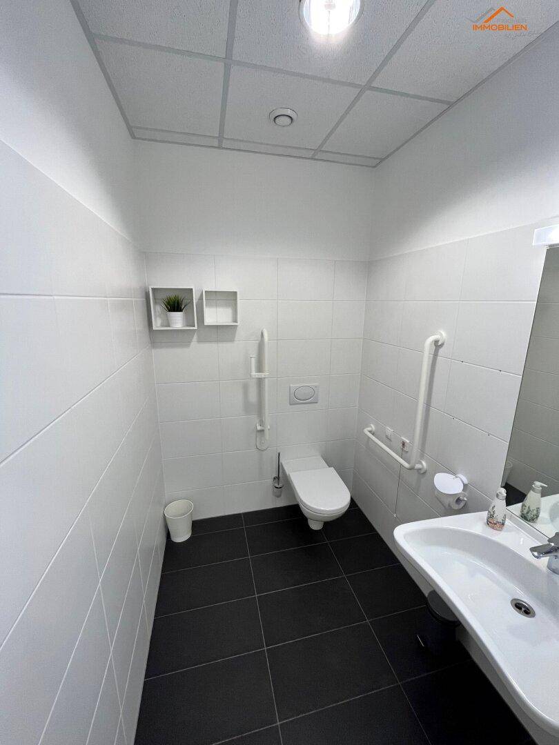 Modernes WC mit weißen Fliesen, dunklem Boden und Haltegriffen für Barrierefreiheit.