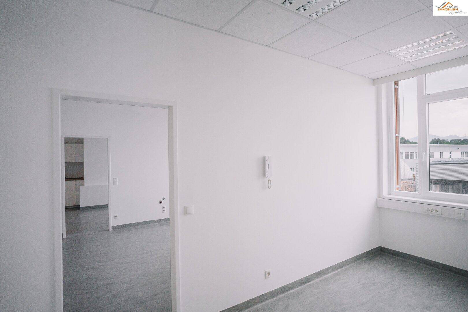Leerer Büroraum mit großem Fenster und Blick in einen angrenzenden Raum.
