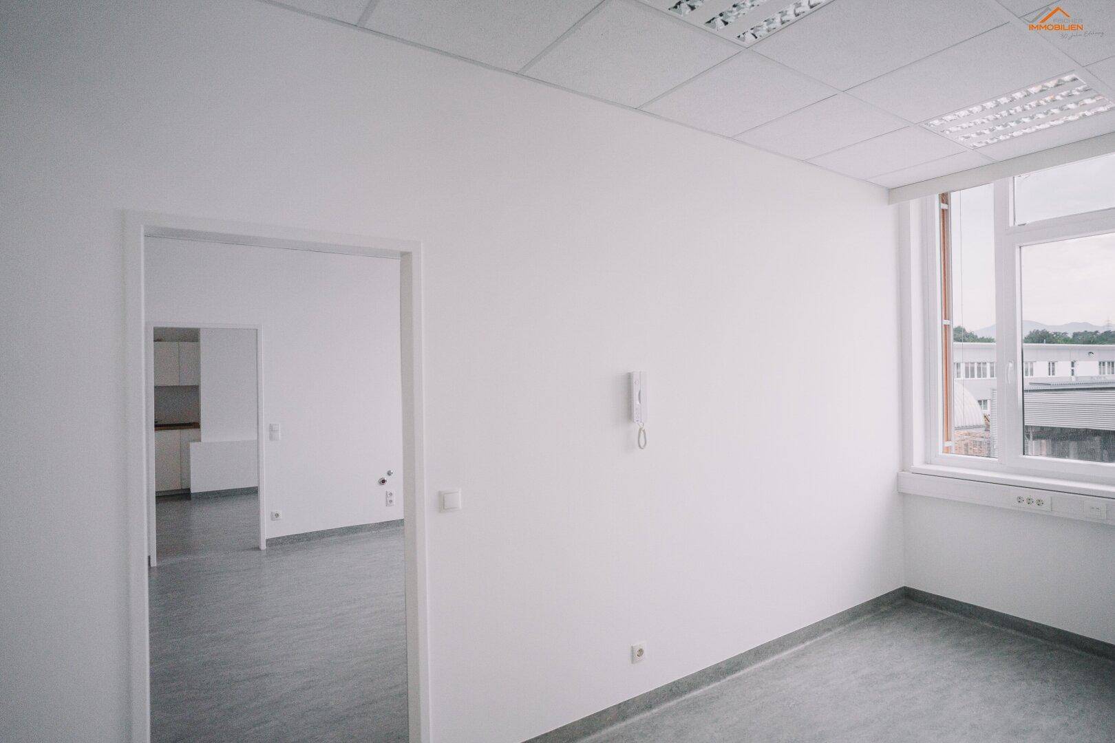 Leerer Büroraum mit Fenster und Blick auf eine angrenzende Küchenzeile.
