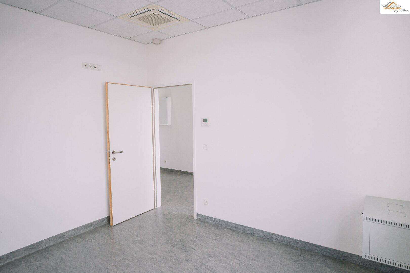 Leerer, heller Büroraum mit Tür, Heizkörper und Vinylboden.