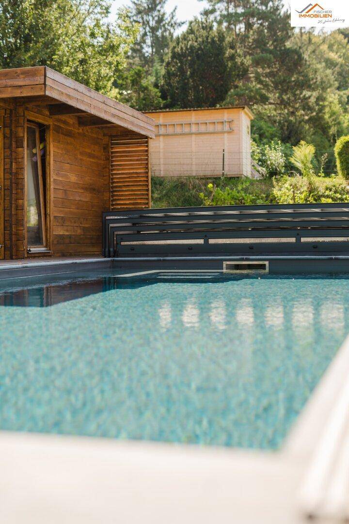 Moderner Poolbereich mit angrenzendem Holzhaus und üppiger grüner Vegetation im Hintergrund.