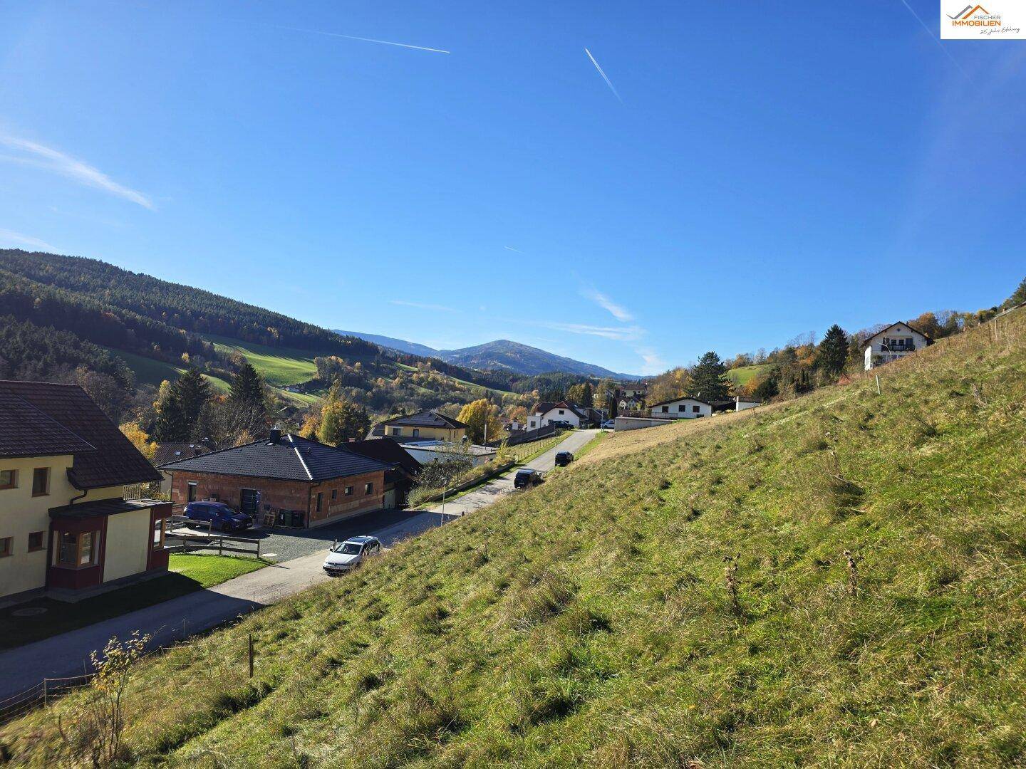 Blick über eine ländliche Siedlung mit Häusern und einer Straße, umgeben von grünen Hügeln und Wäldern.