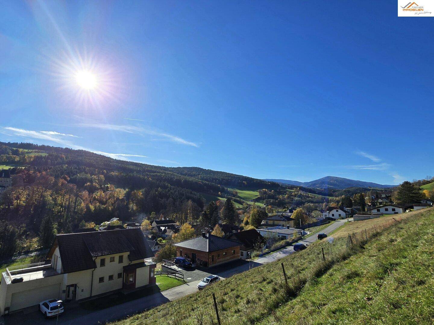Weitläufiger Blick über eine Siedlung in hügeliger Landschaft mit Wald und klarem blauen Himmel.