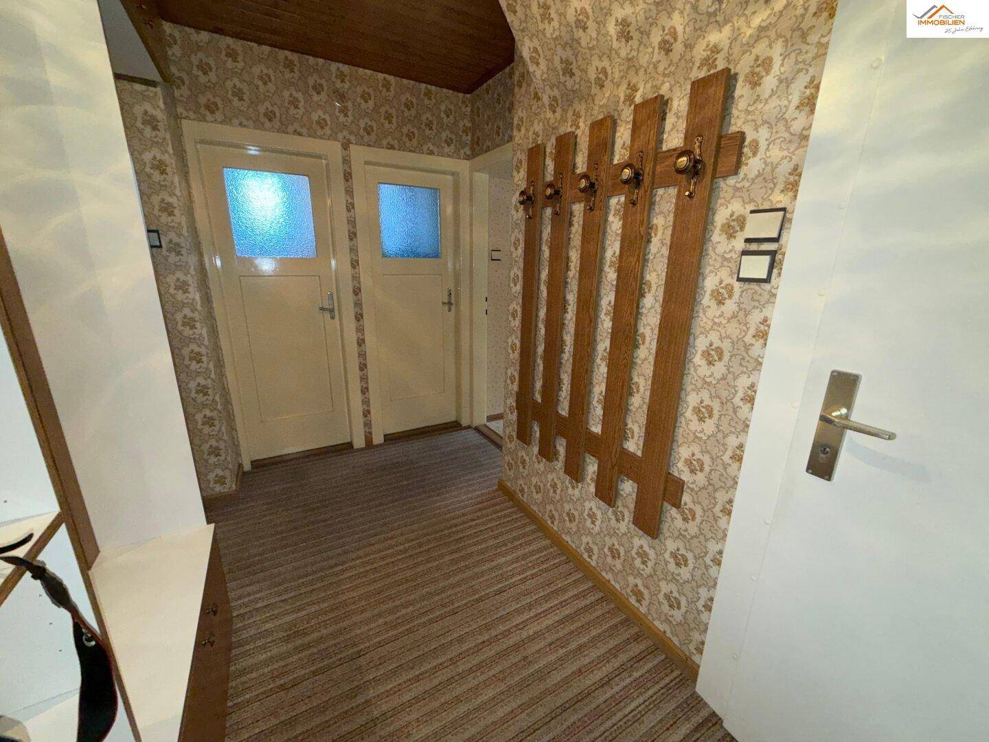 Flur mit gemusterter Tapete, Holzdecke und Garderobe, der zu weiteren Räumen führt.