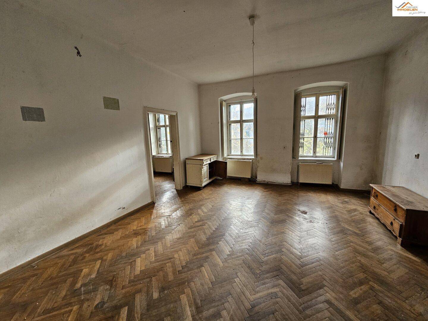 Helles Zimmer mit Fischgrätparkett und zwei großen Fenstern, die viel Tageslicht hereinlassen.