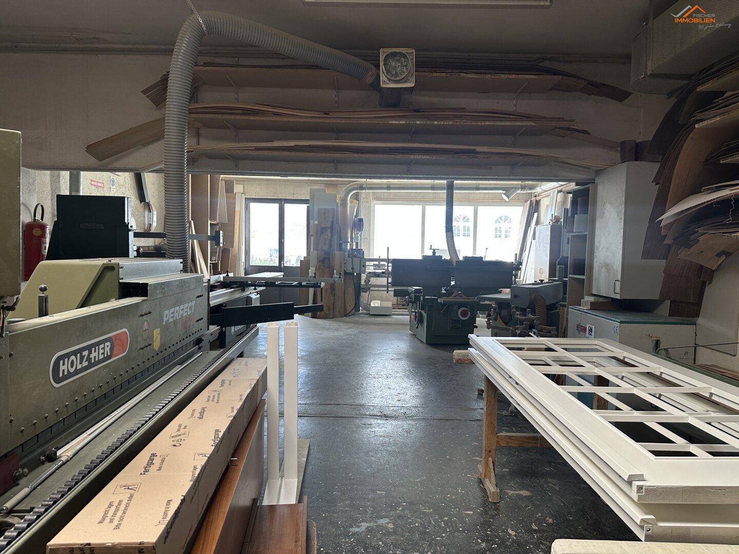 Innenansicht einer Holzwerkstatt mit professionellen Maschinen, Holzplatten und viel Tageslicht durch große Fenster.