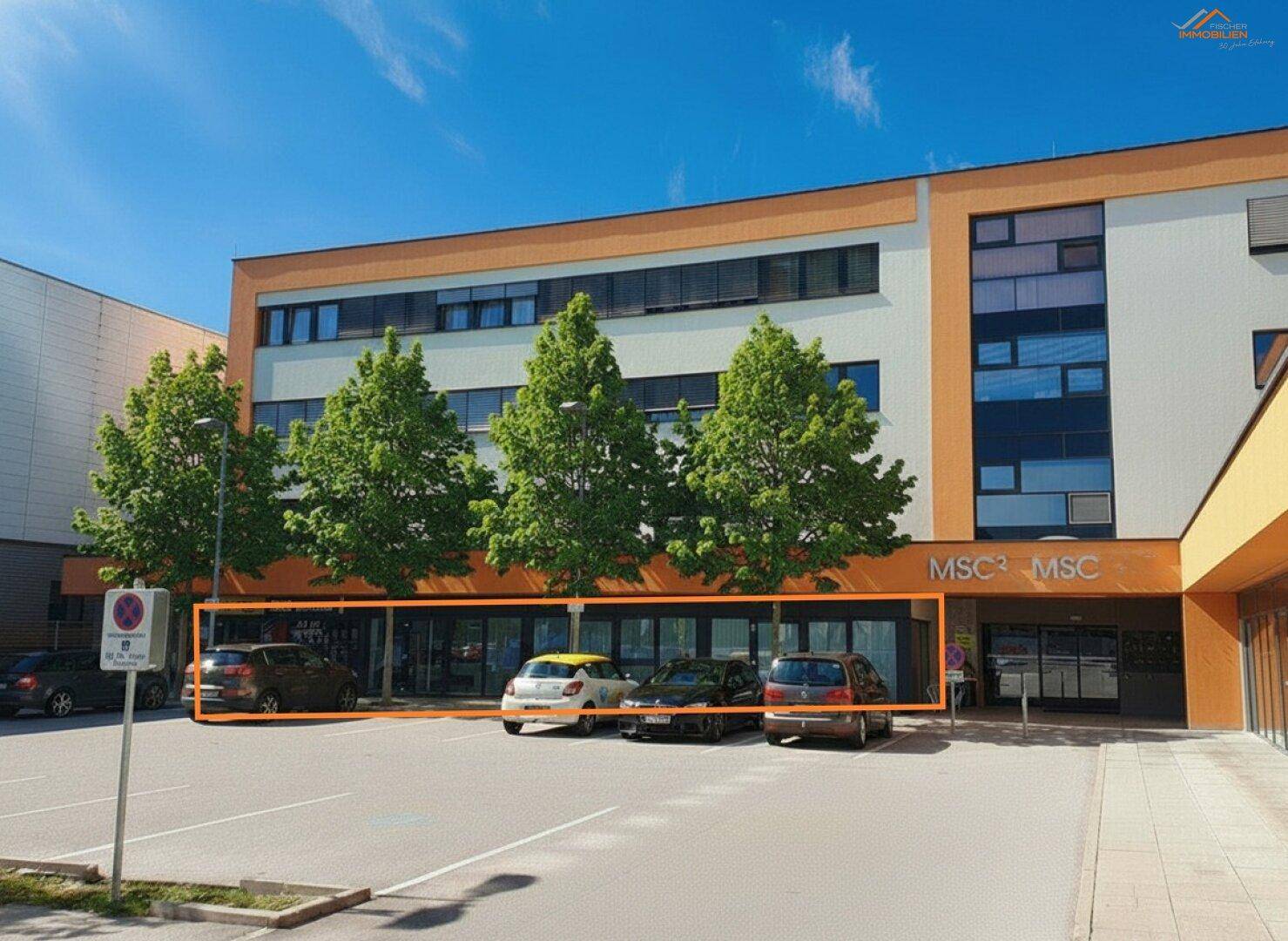 Außenansicht des Geschäftsgebäudes mit Parkplatz und hervorgehobenem Erdgeschossbereich unter Bäumen.