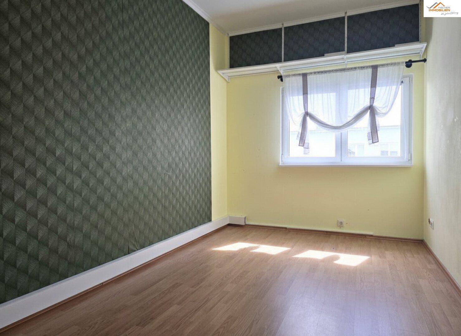 Leerer Raum mit Laminatboden und einer Wand mit geometrischer Tapete, gut beleuchtet durch ein Fenster.