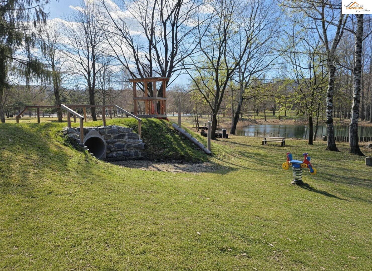 Spielbereich mit Rutsche, Kletterturm und einem kleinen Teich im Hintergrund.