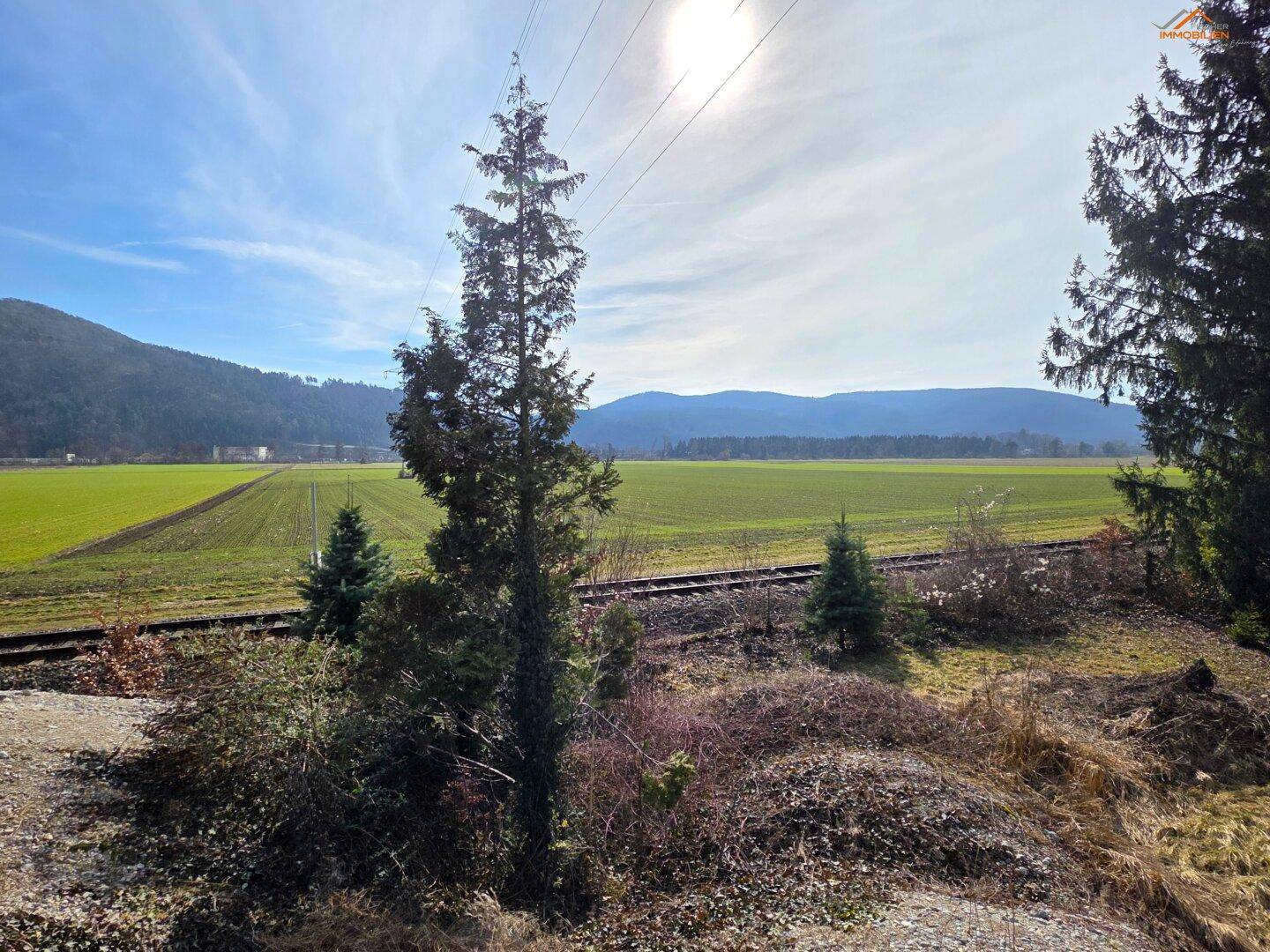 Weitläufige grüne Felder mit Bahngleisen im Vordergrund und einer Bergkette am Horizont.