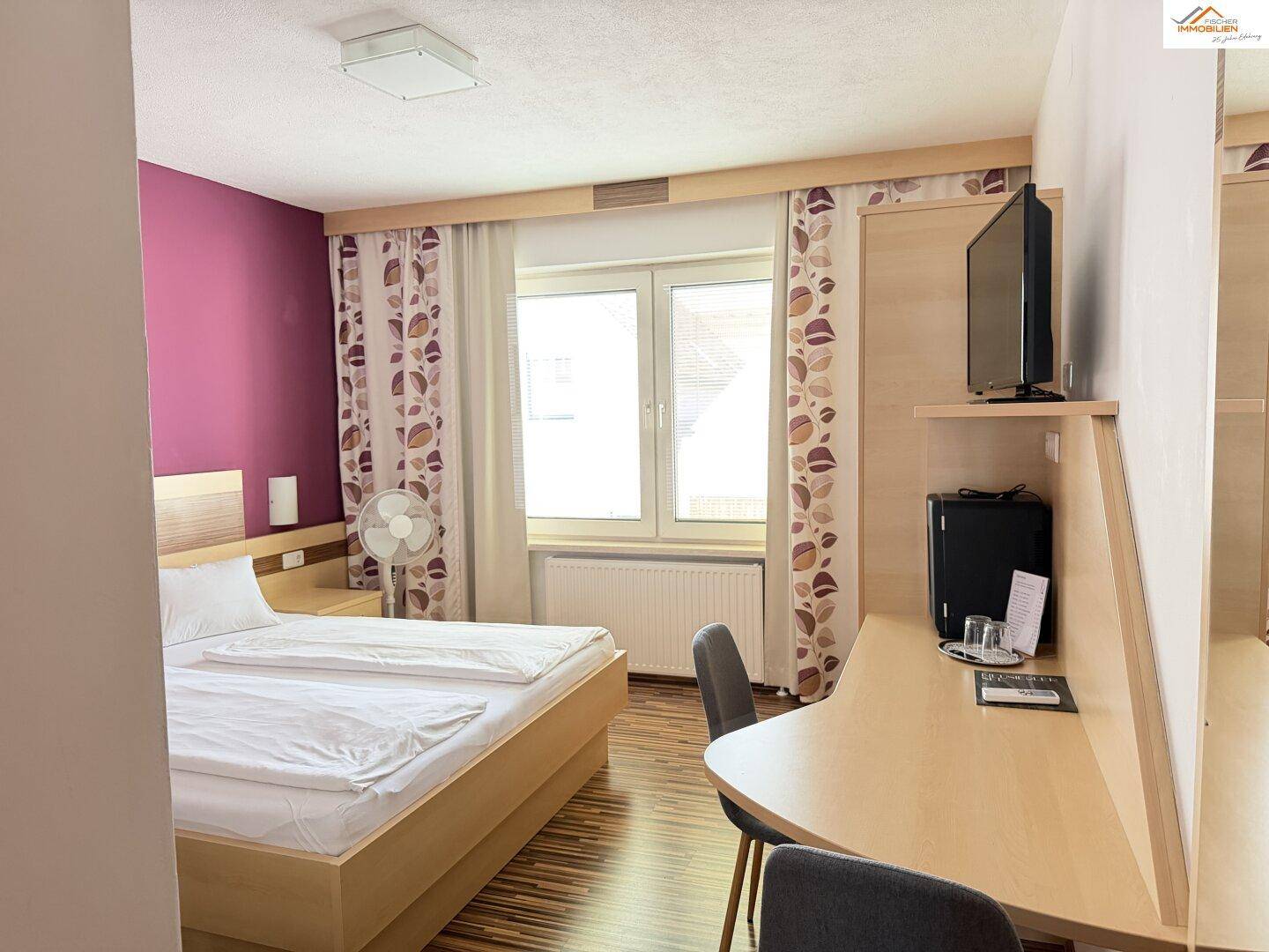 Helles Schlafzimmer mit Doppelbett, Schreibtisch und Fenster mit Vorhängen.