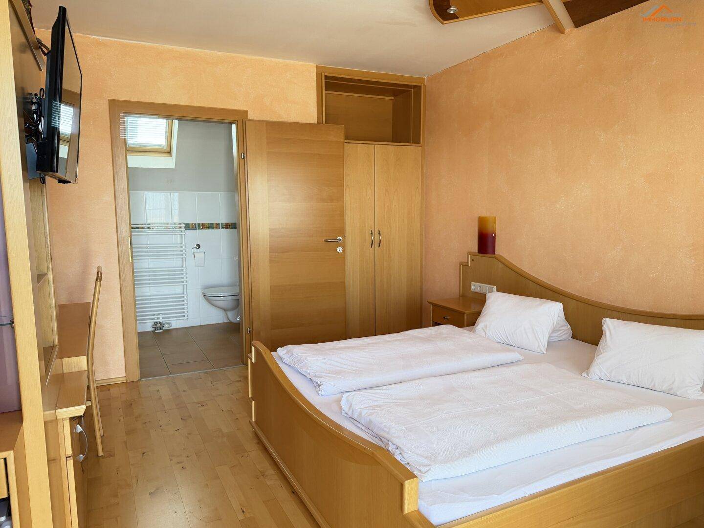 Schlafzimmer mit Doppelbett und Holzmöbeln, das den direkten Zugang zum angrenzenden Bad zeigt.
