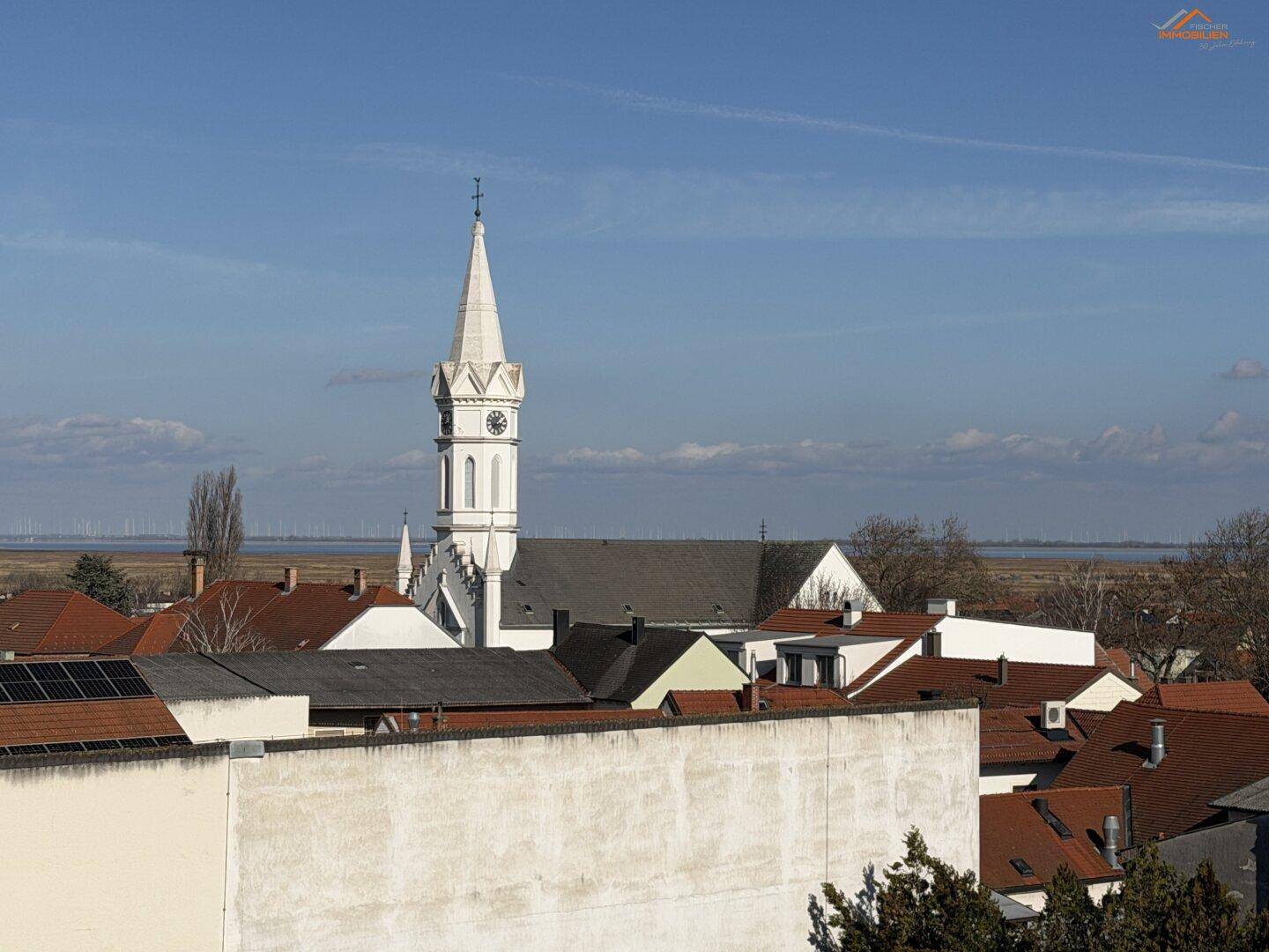 Blick auf einen Kirchturm und die Dächer des Dorfes mit einem See in der Ferne.